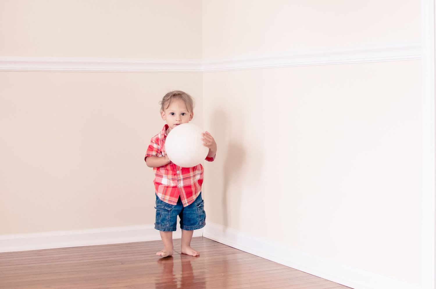 Child in red plaid shirt holding white balloon in empty room corner.