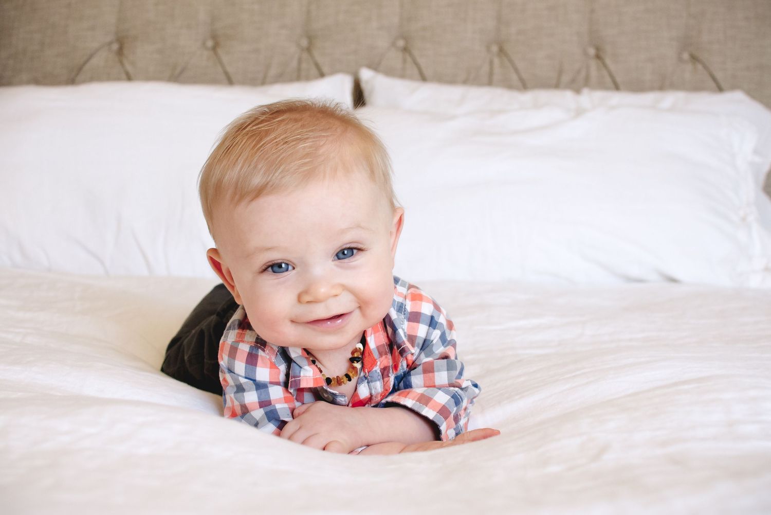 Baby in red plaid shirt lying on white bedding with a bright smile.