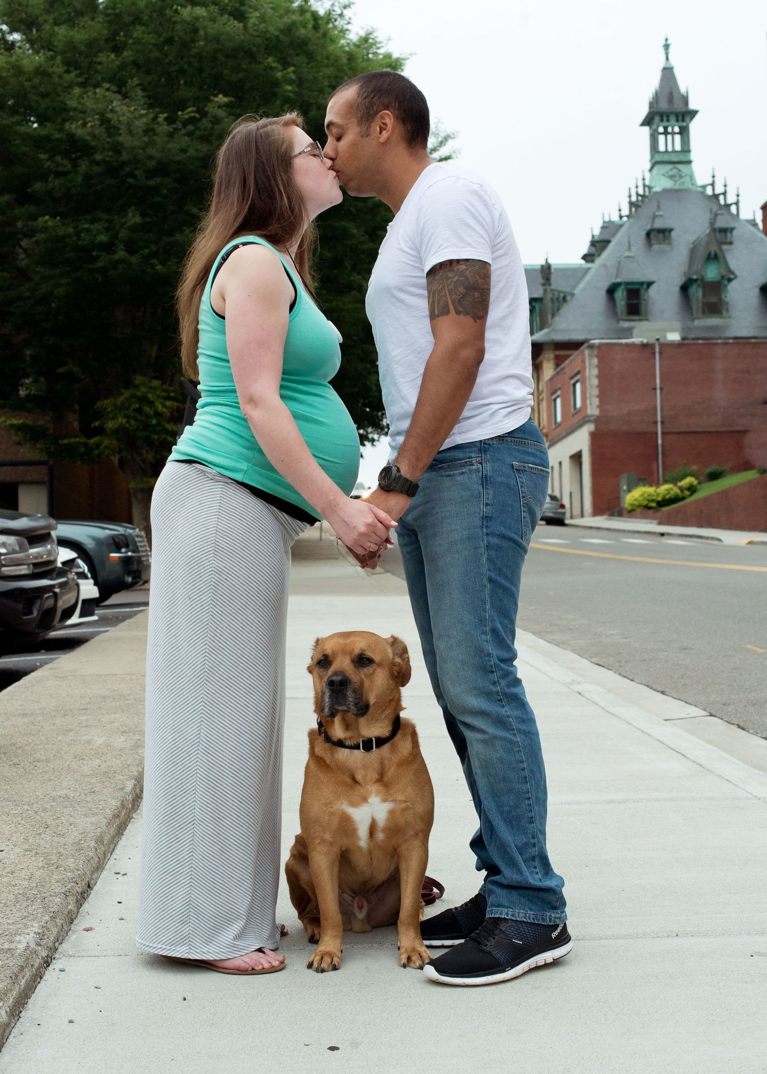 A couple stands together on a sidewalk with their brown dog in a series of romantic engagement photos.