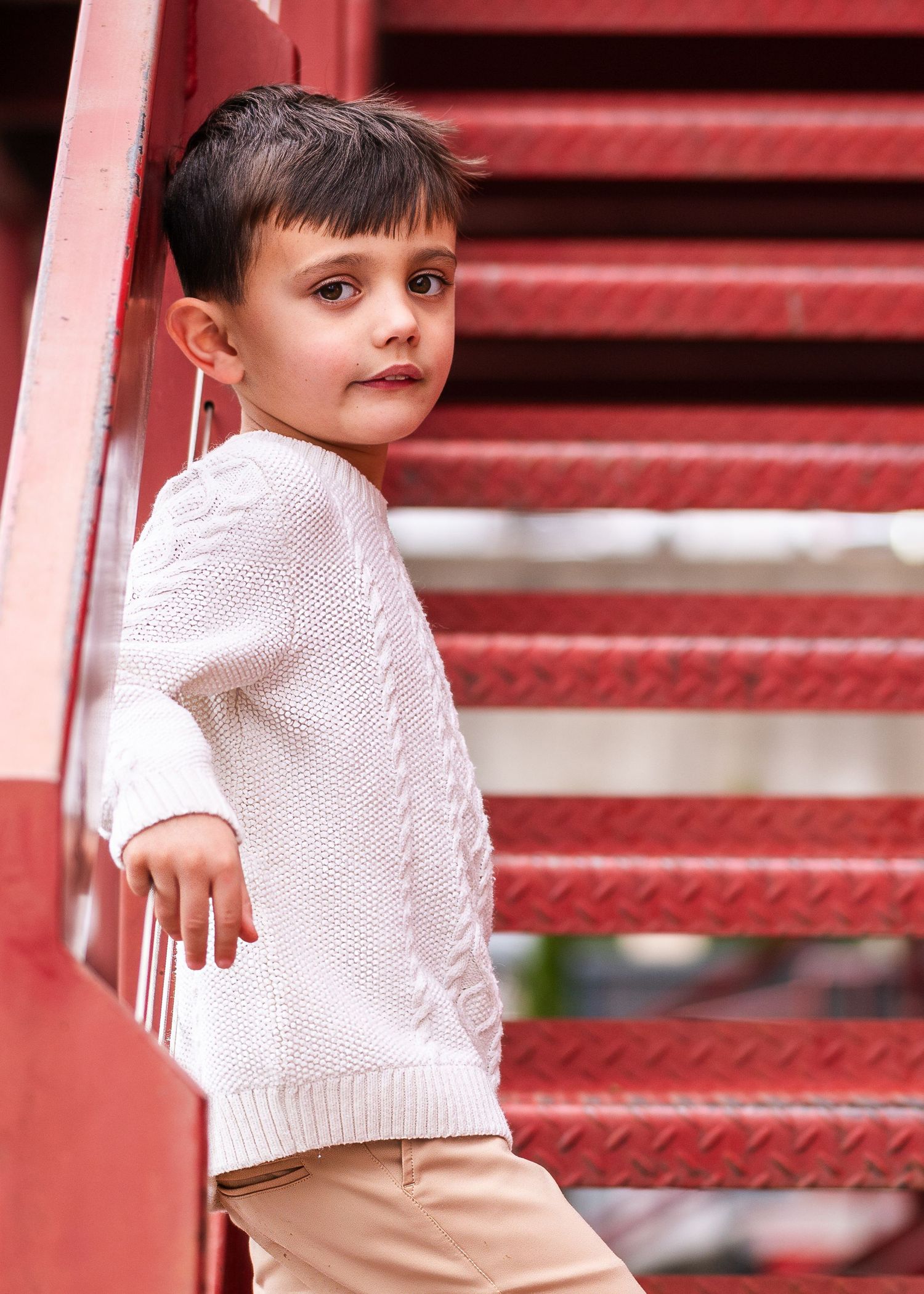 Child in white sweater posing on red metal stairs against brick wall.