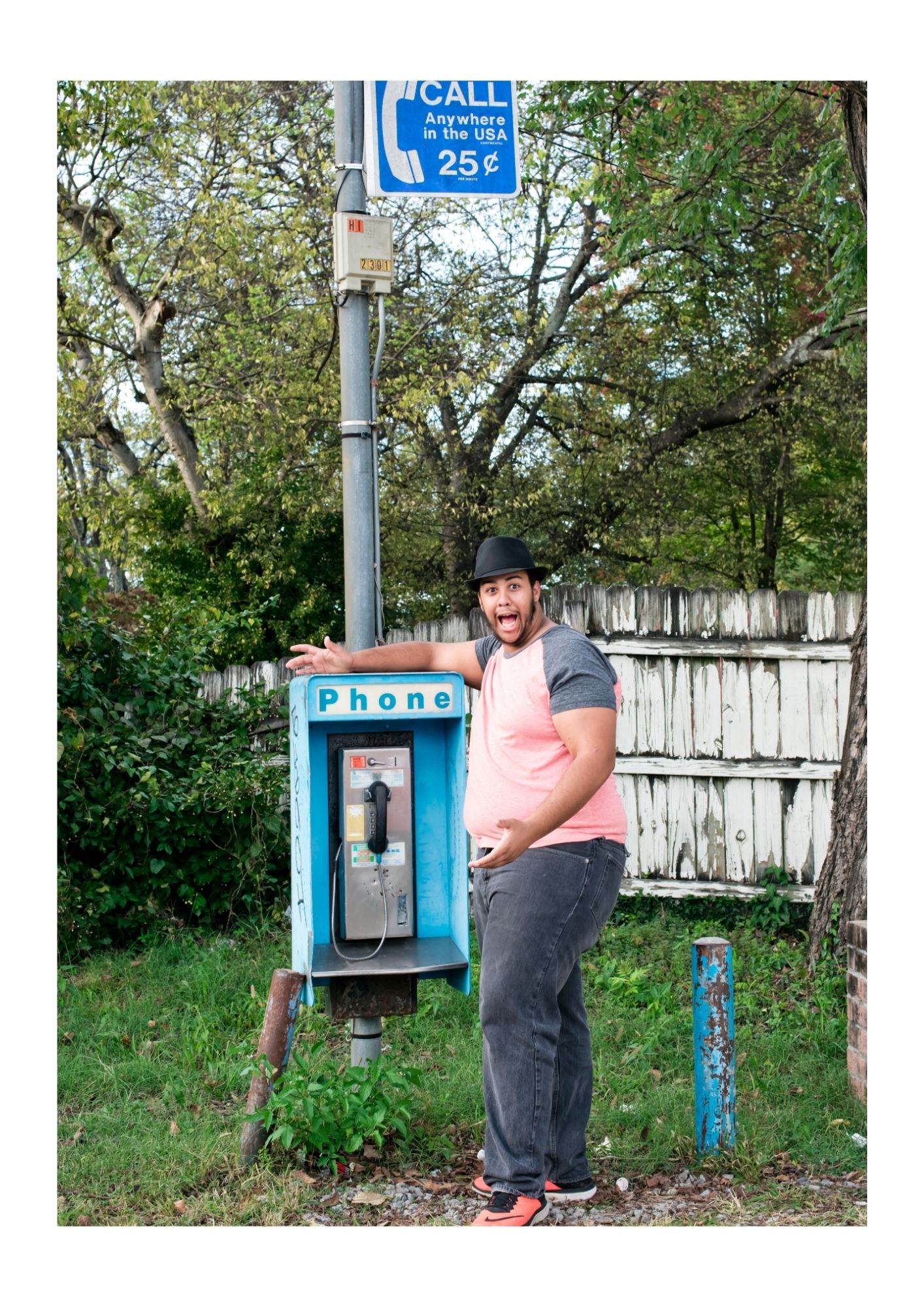 Person posing playfully next to vintage blue payphone booth in outdoor setting. Senior portrait