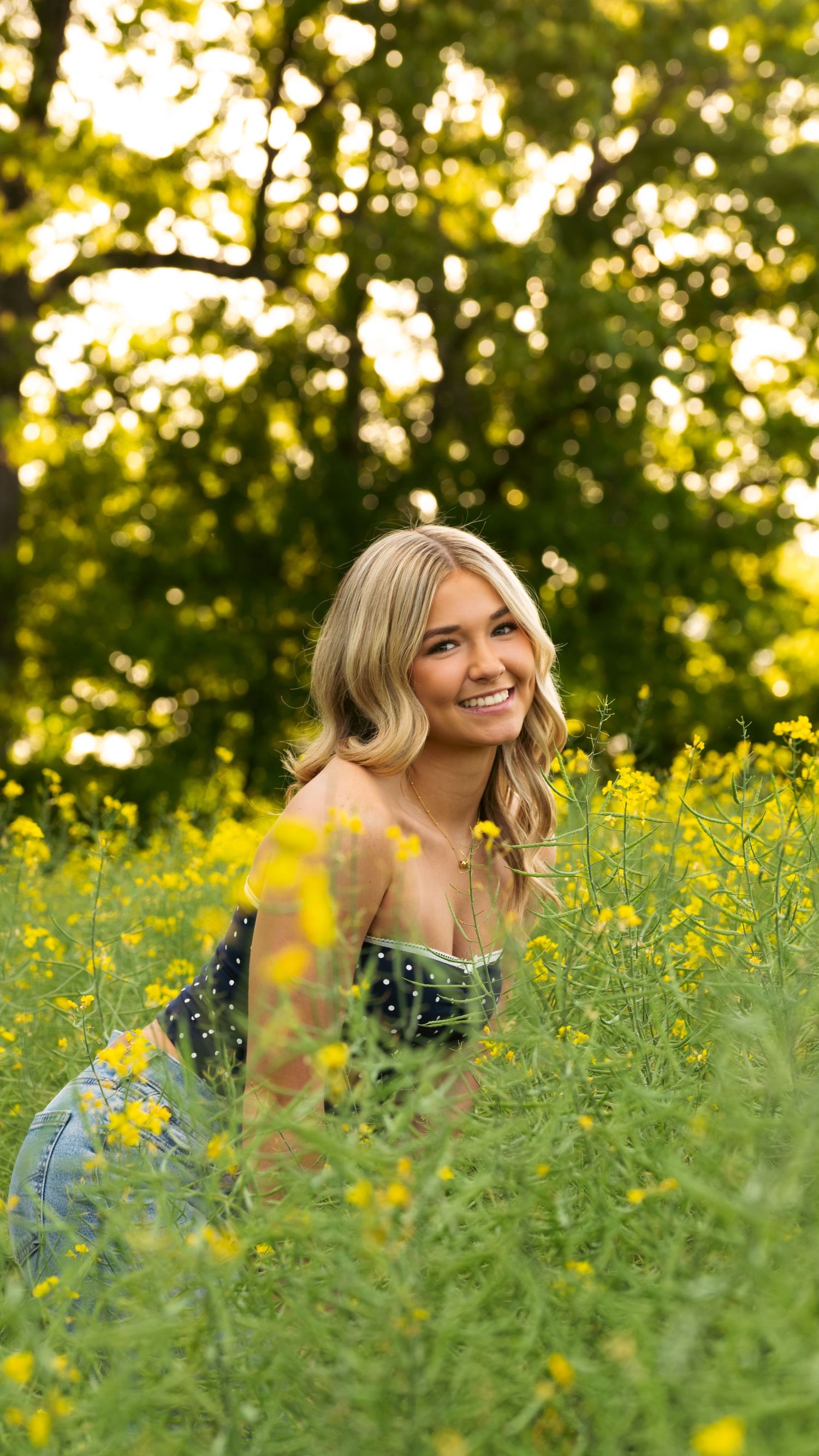 Smiling blonde woman sitting in a field of yellow wildflowers with lush green trees in the background.