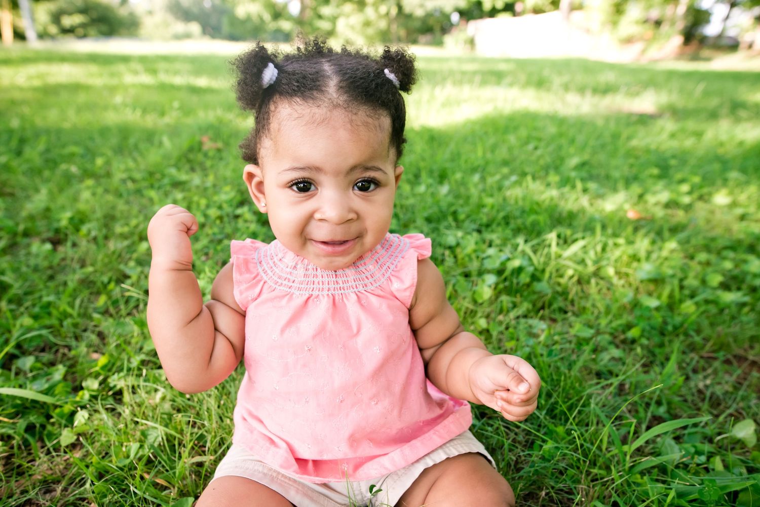 A small child in a pink dress sits and plays on lush green grass during a sunny summer day.