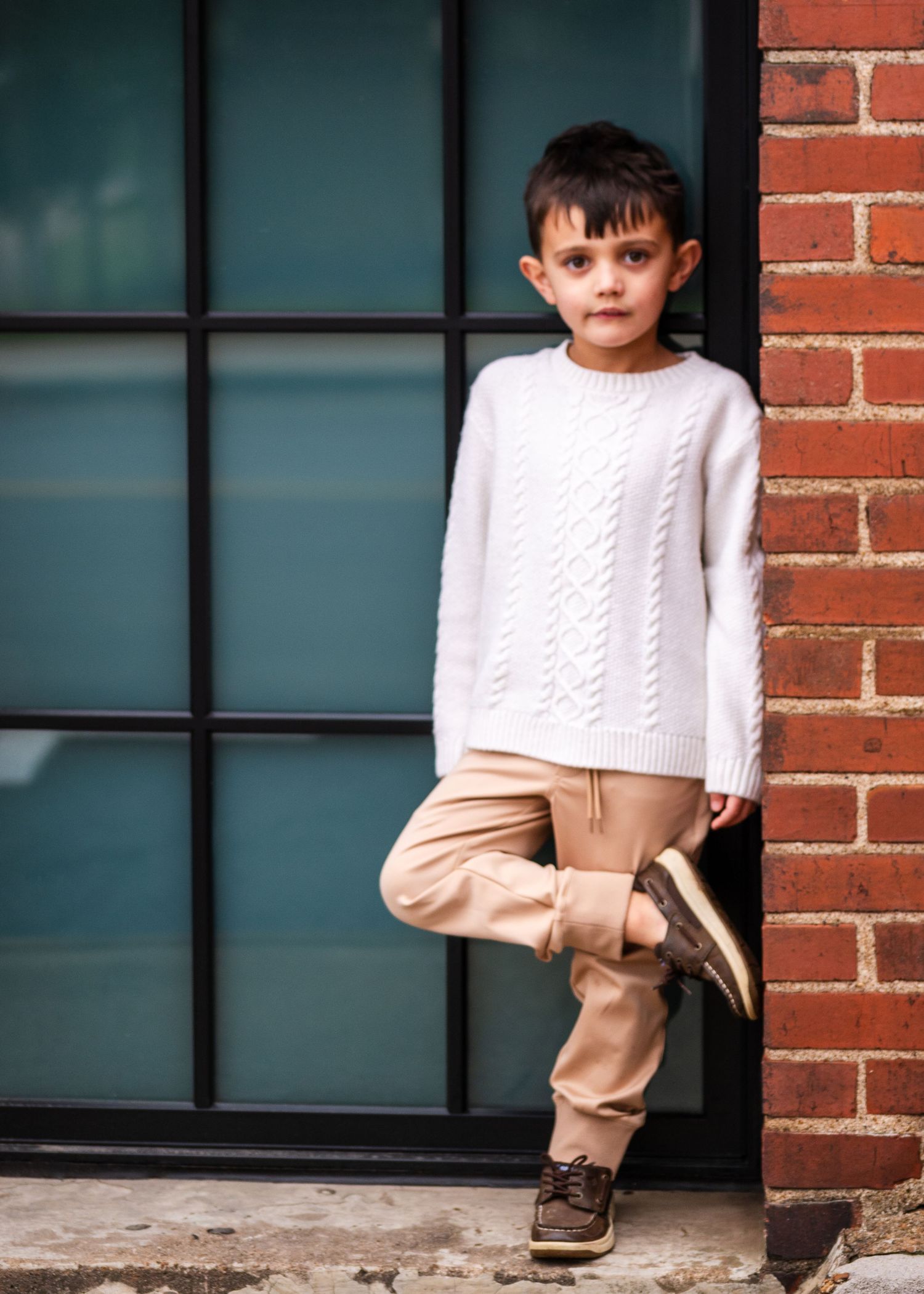 Child in white cable knit sweater and beige pants leaning against brick wall with window.