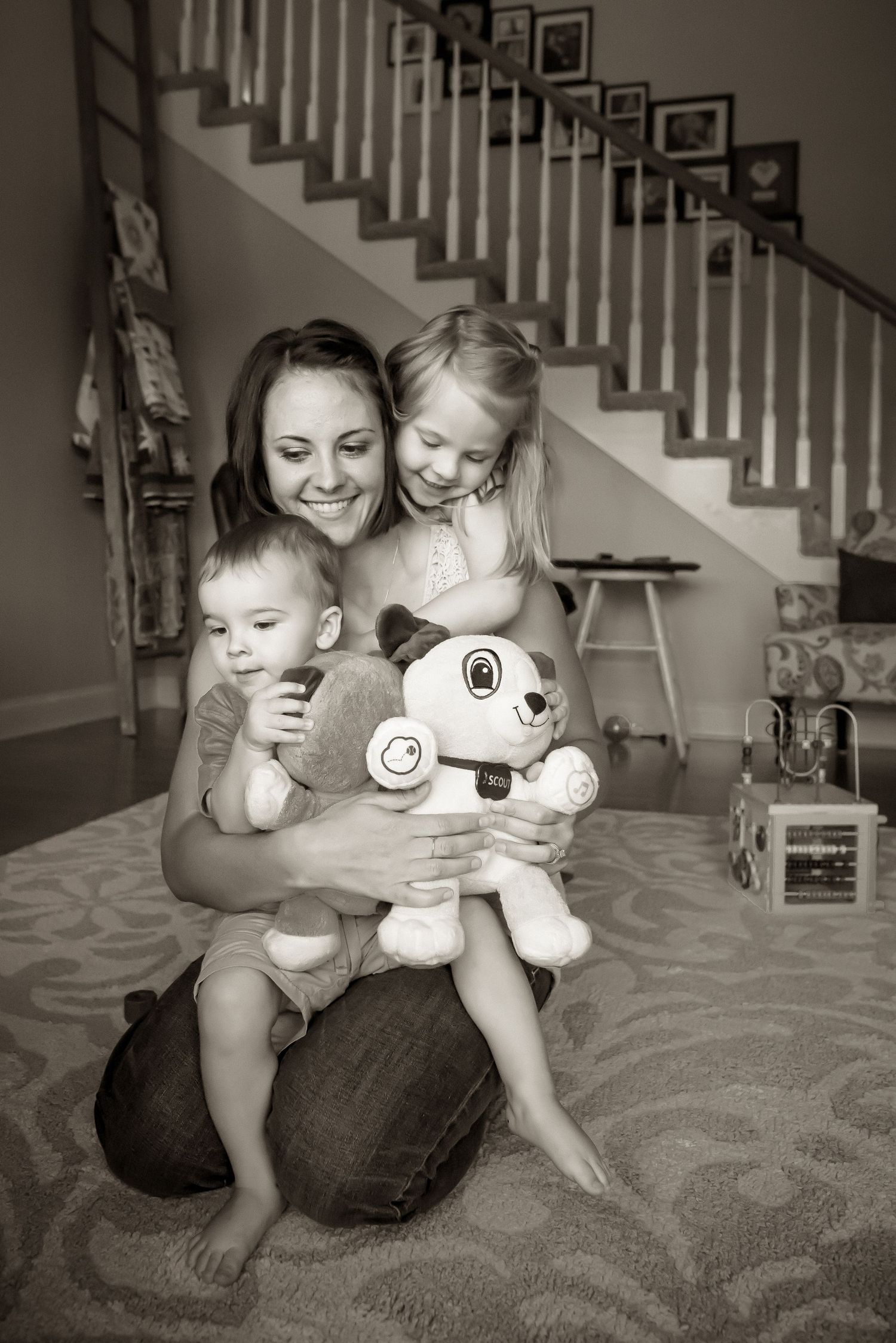 Black and white photo of family moment with stuffed toy on carpeted floor by staircase.