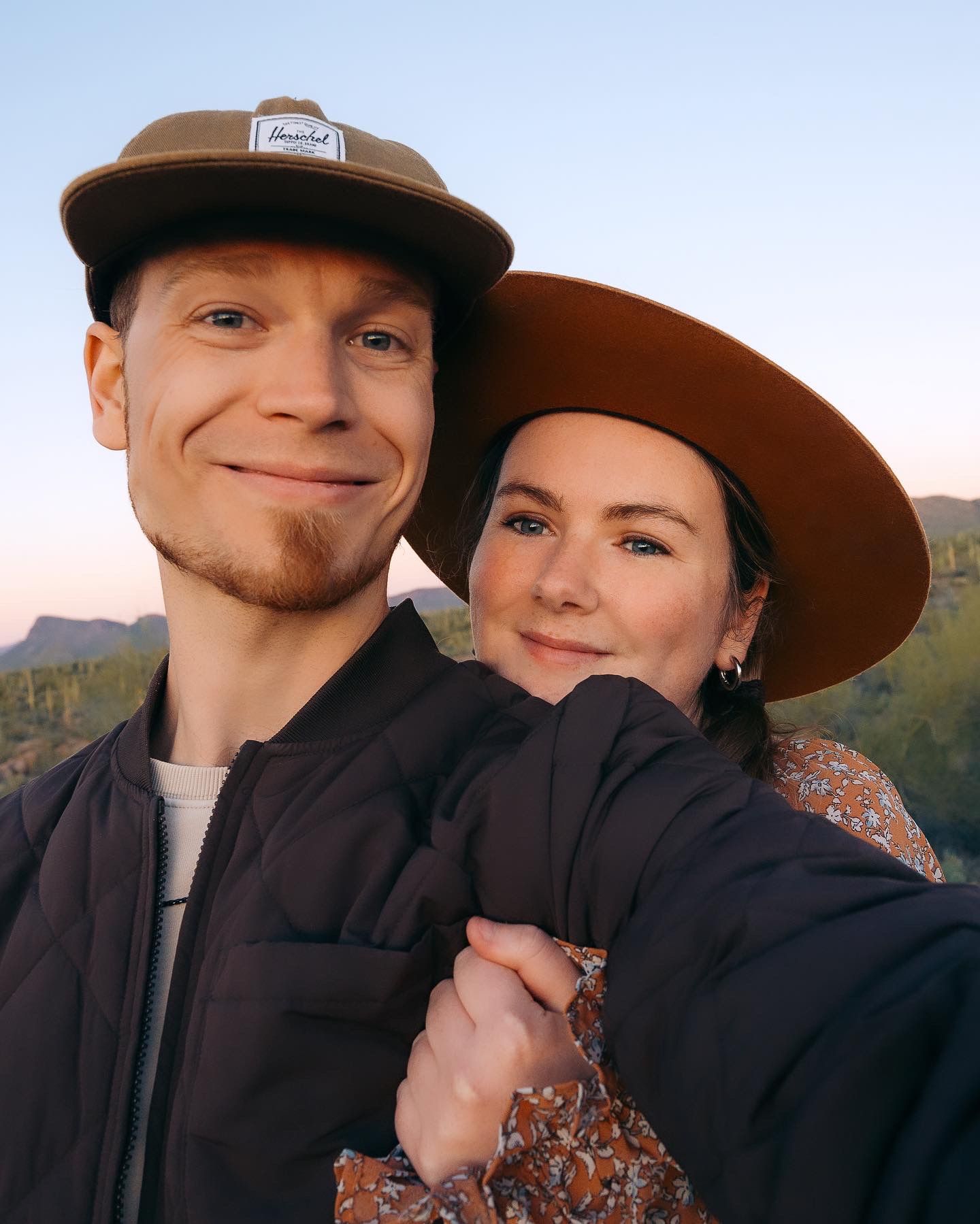Smiling couple in hats taking a selfie with a scenic desert mountain sunset backdrop.