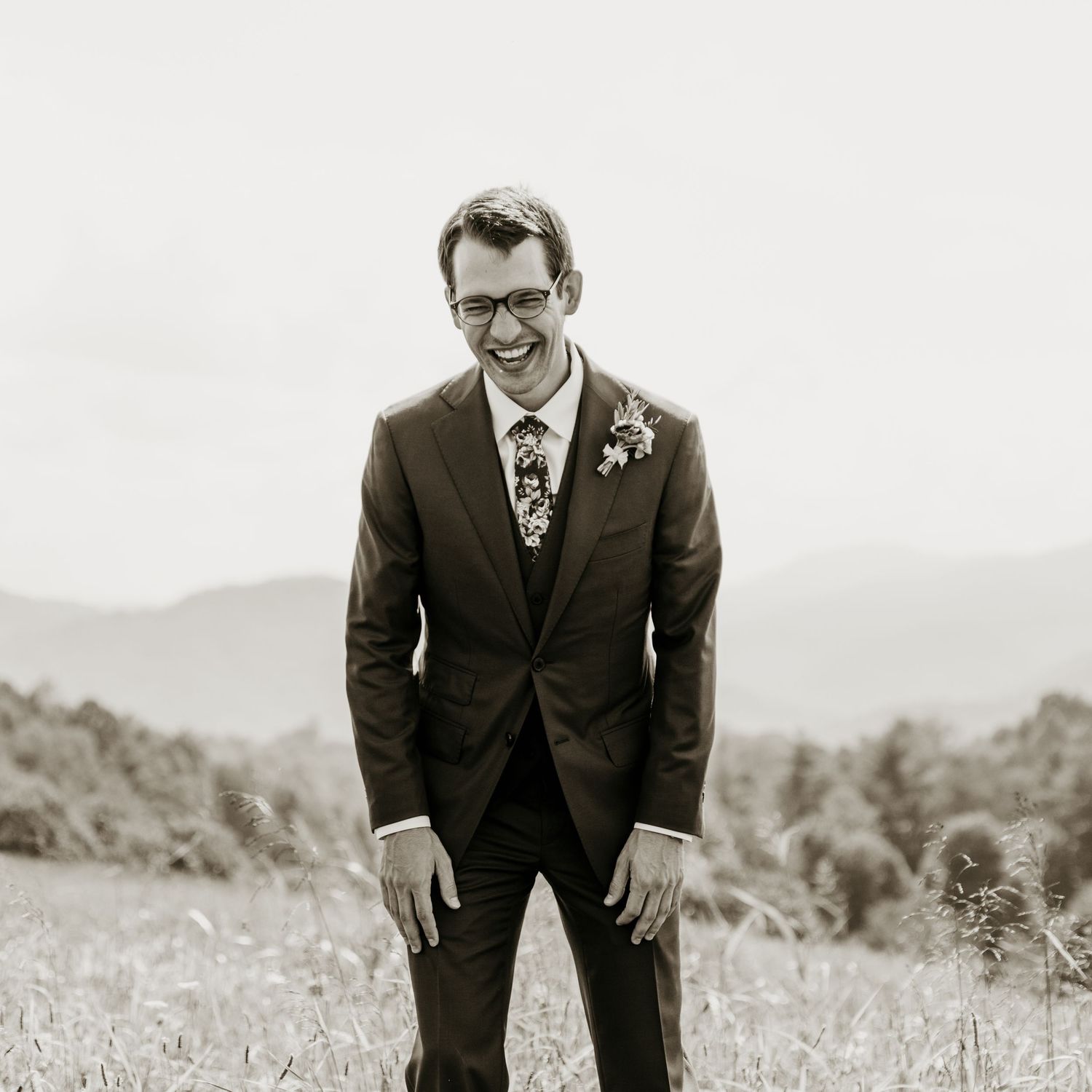 Smiling groom in dark suit with floral tie stands in a field with mountain backdrop in black and white photo.