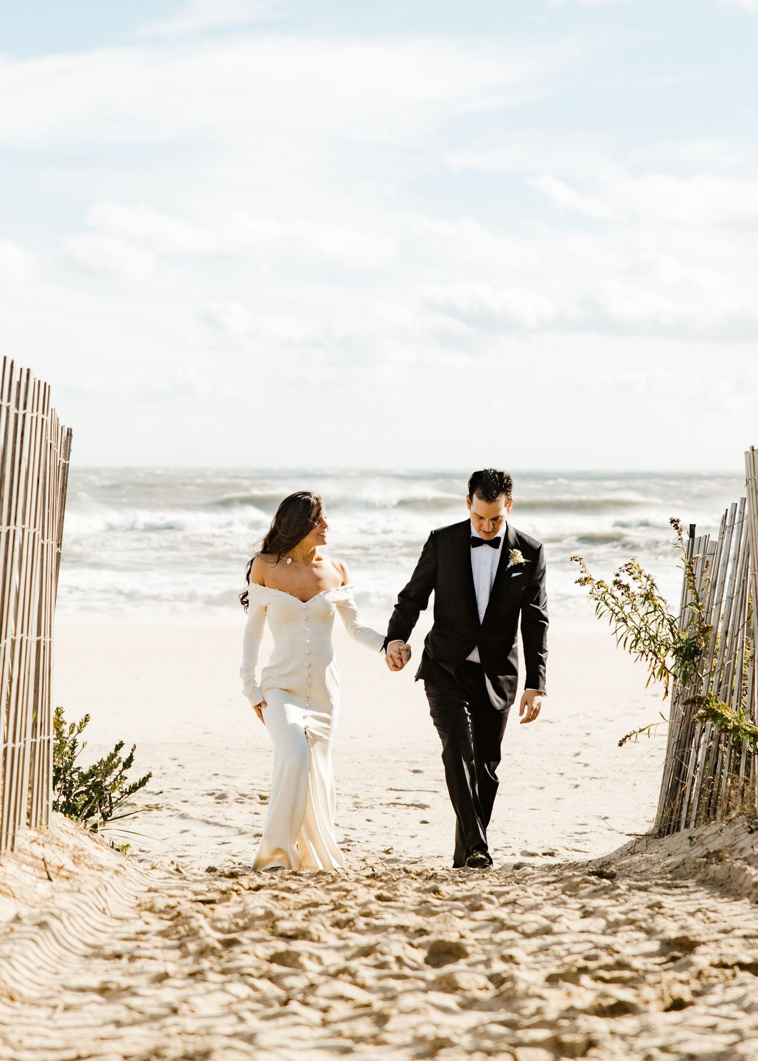 Bride and groom walking hand in hand through a beach pathway lined with wooden fences.