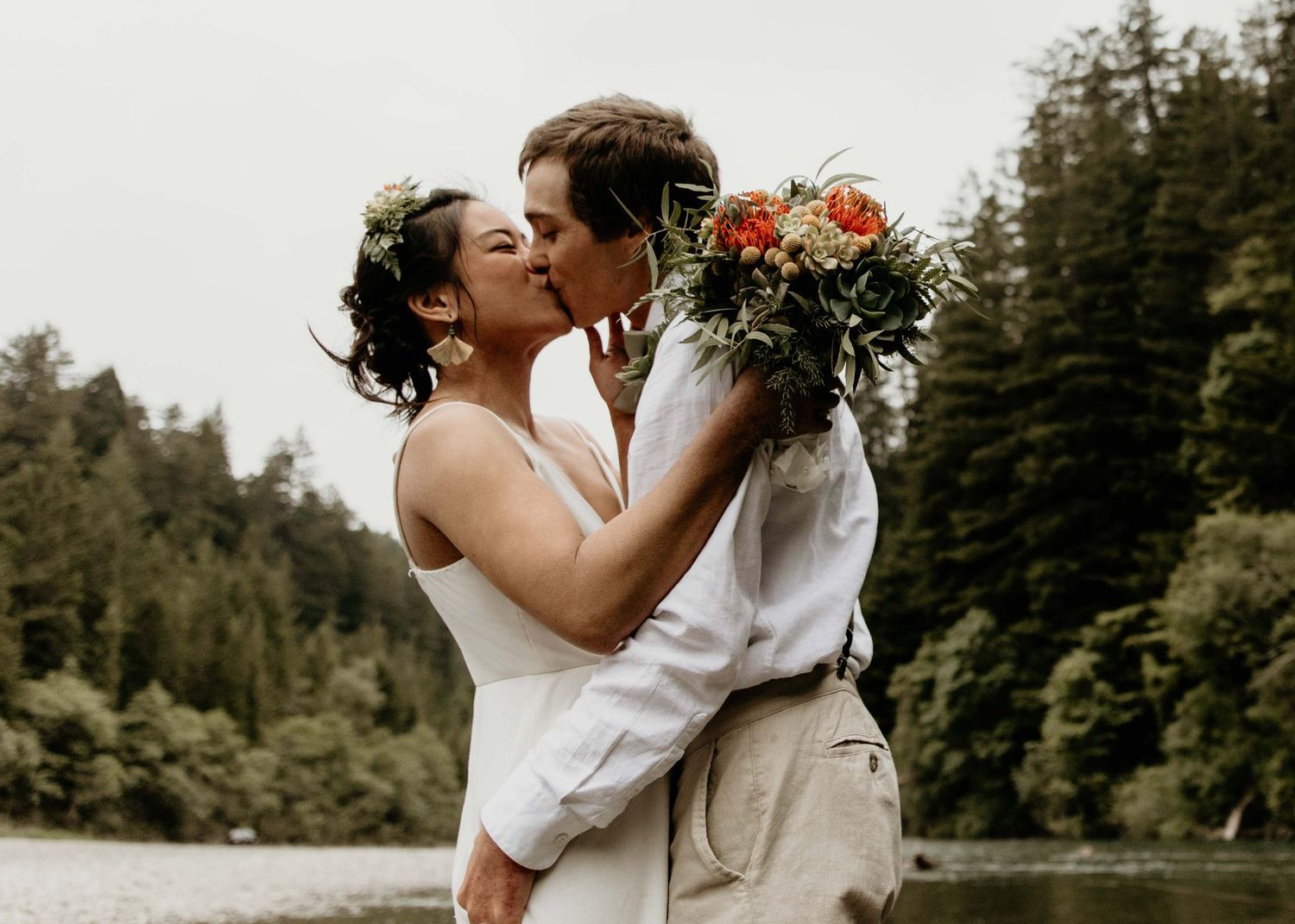 Two brides sharing a passionate kiss outdoors by a river, one holding a vibrant orange and green bouquet.