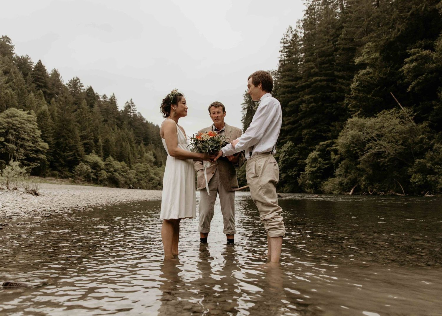 Bride and groom exchanging vows with officiant while standing in a shallow forest river.