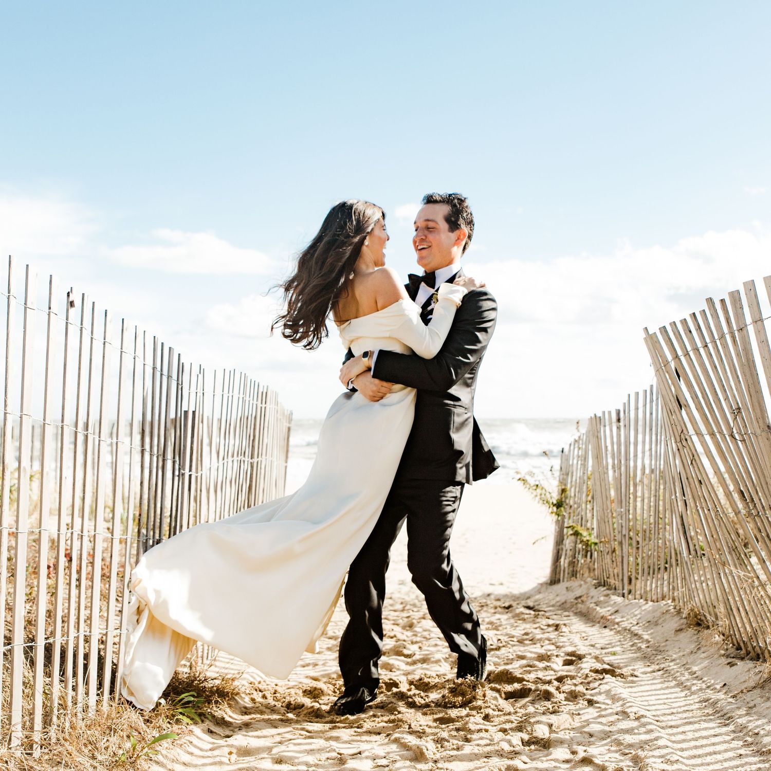 Groom lifts his bride in a flowing white gown on a sandy beach path lined with wooden fences.