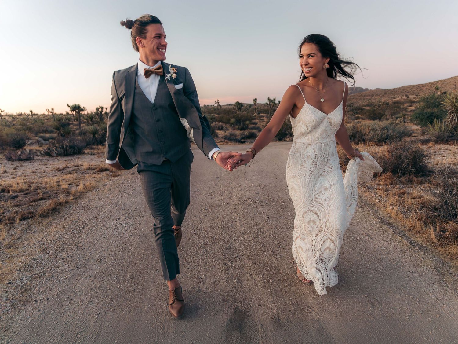 Bride and groom walking hand in hand down desert road at sunset.