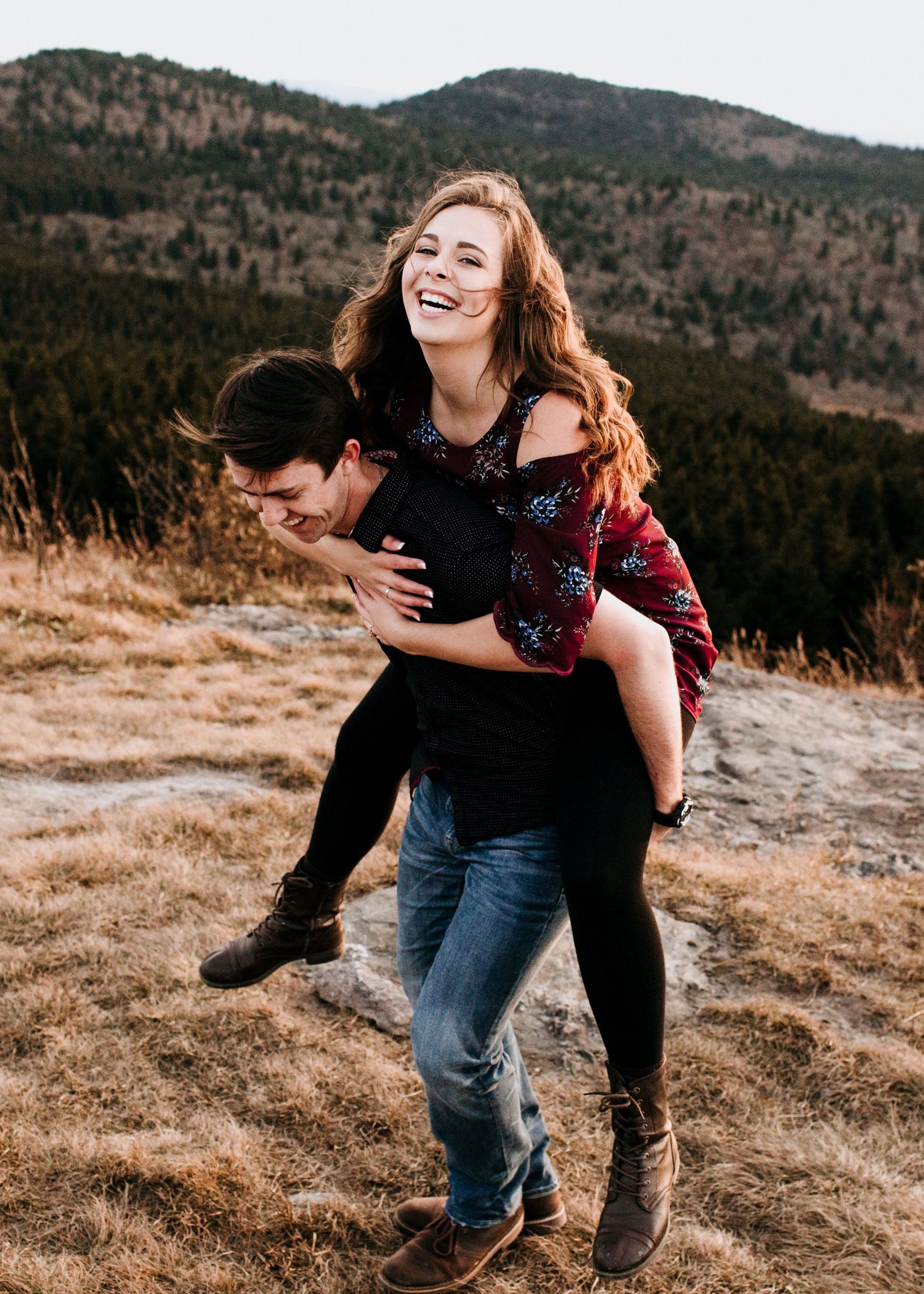 Happy couple laughing as man gives woman a piggyback ride on a mountain trail during engagement photos.