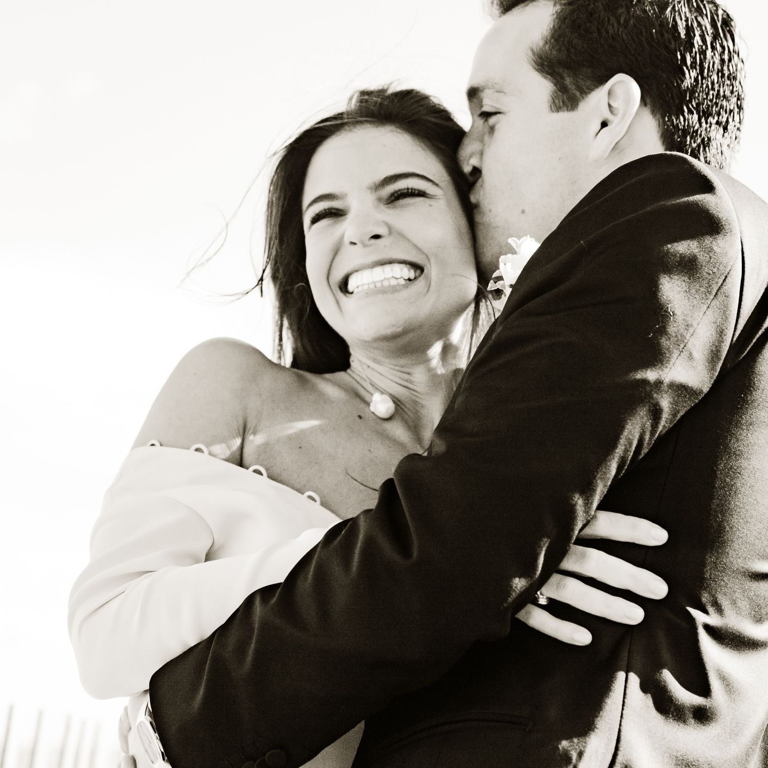 Black and white photo of a smiling bride being embraced and kissed by her groom on their wedding day.