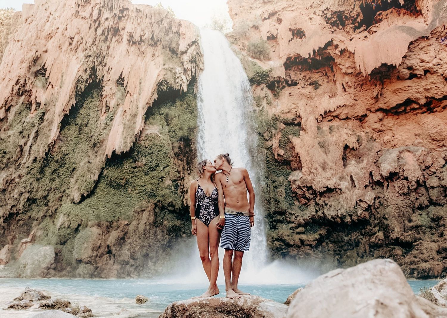 Couple standing on a rock kissing in front of a stunning turquoise waterfall canyon.