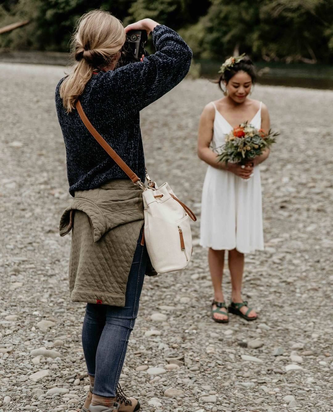 Photographer capturing a bride holding a bouquet on a rocky riverbank during an outdoor wedding shoot.
