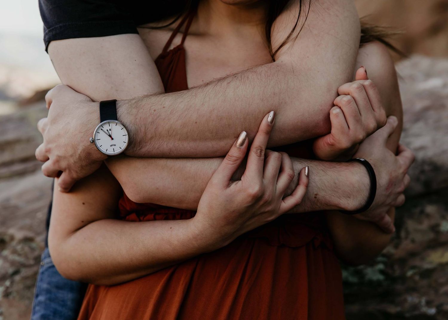 Couple embracing tenderly, intertwined arms showing a watch and manicured nails against a rustic outdoor backdrop.