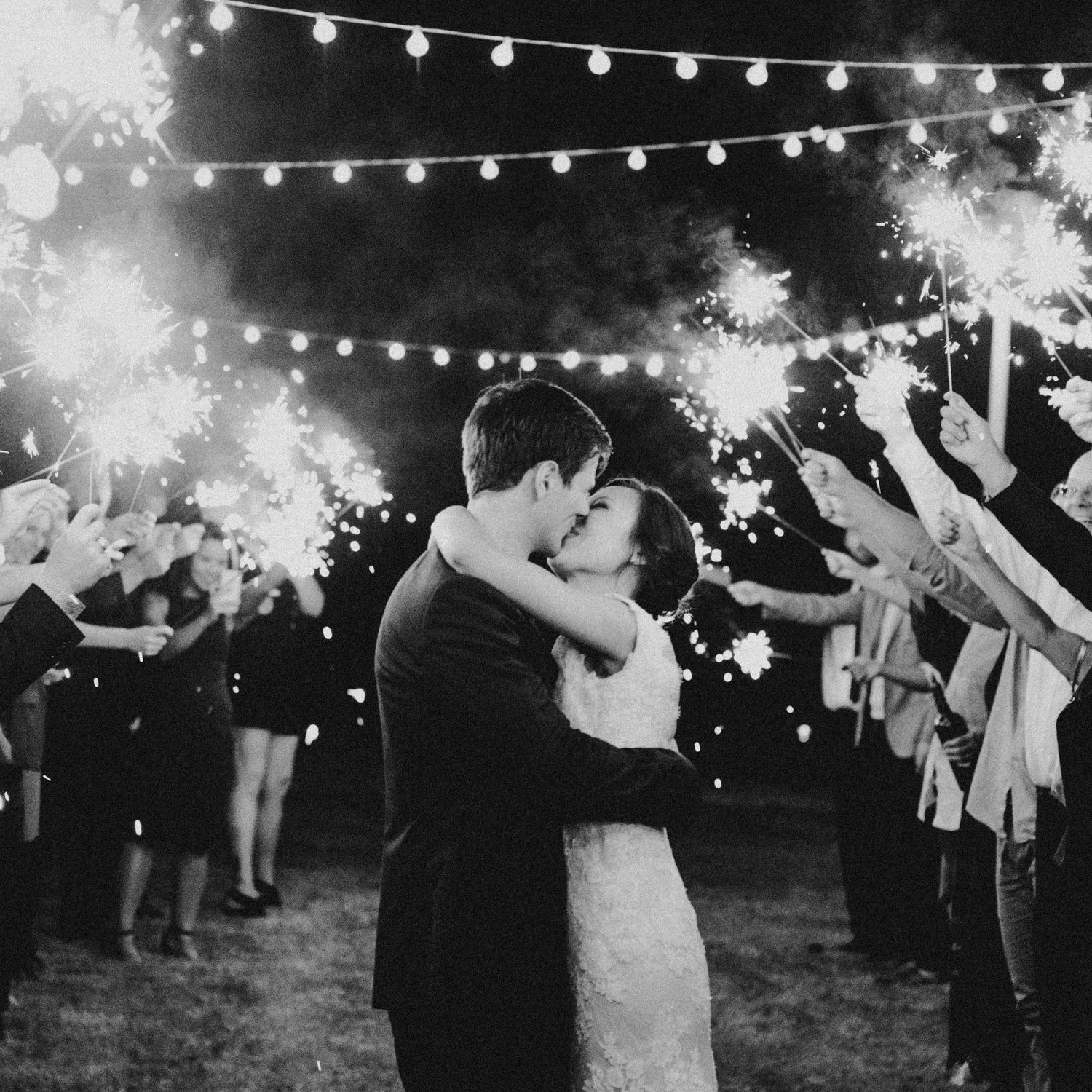 Black and white photo of newlyweds kissing during a romantic sparkler send-off under string lights at night.