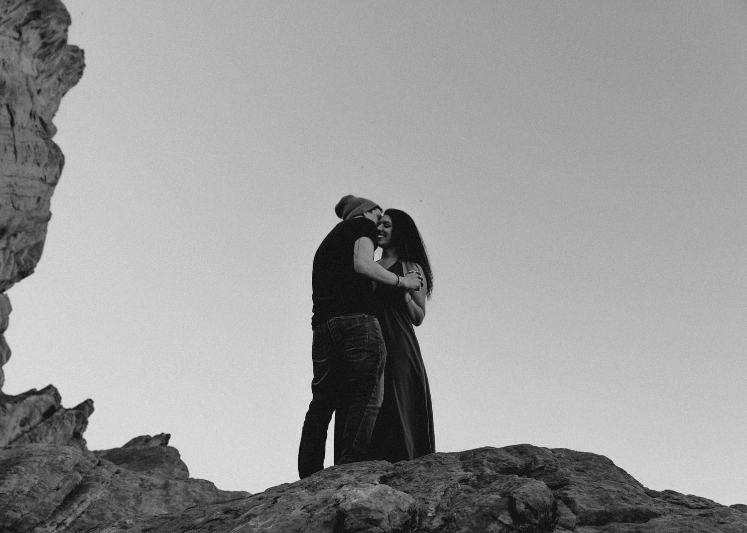 Black and white photo of a couple embracing and kissing on a rocky hilltop against a clear sky.