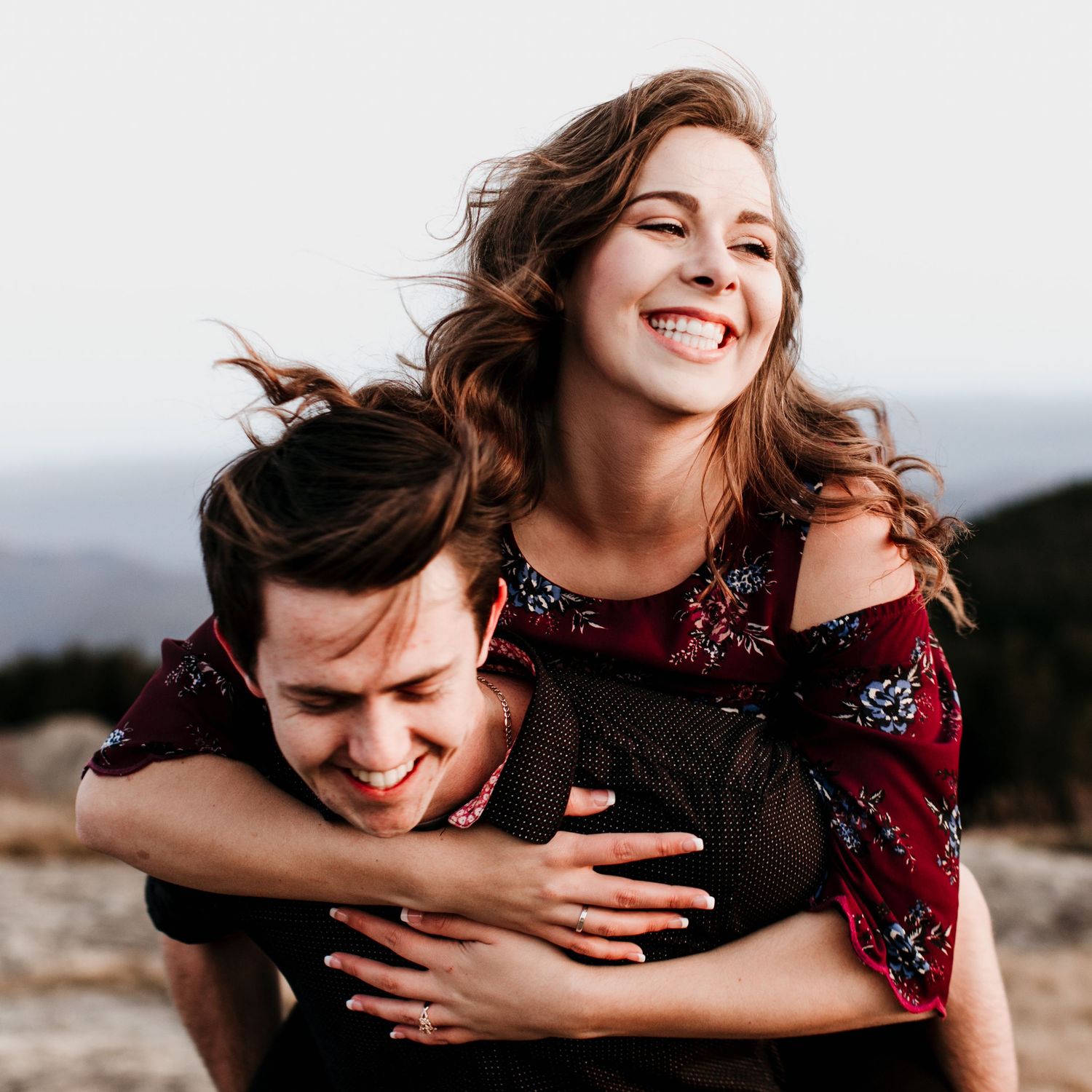 Joyful couple laughing together as woman rides piggyback on mans back against a scenic sky backdrop.
