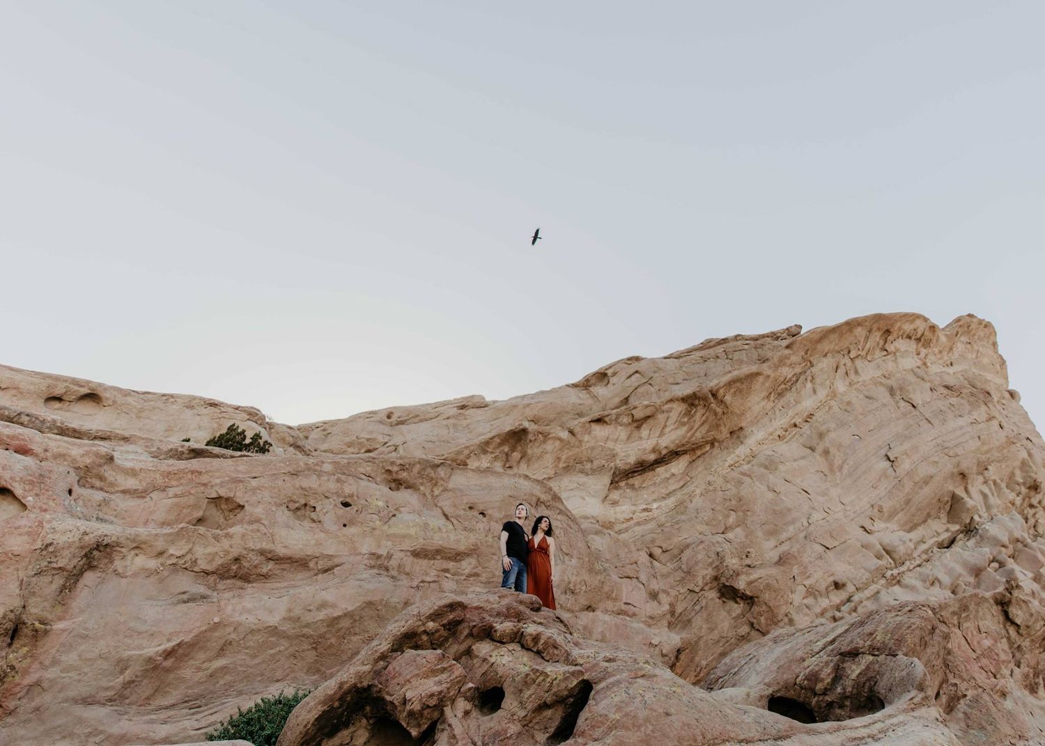 Couple standing together on dramatic rocky desert cliffs with a vast open sky and a bird soaring above.