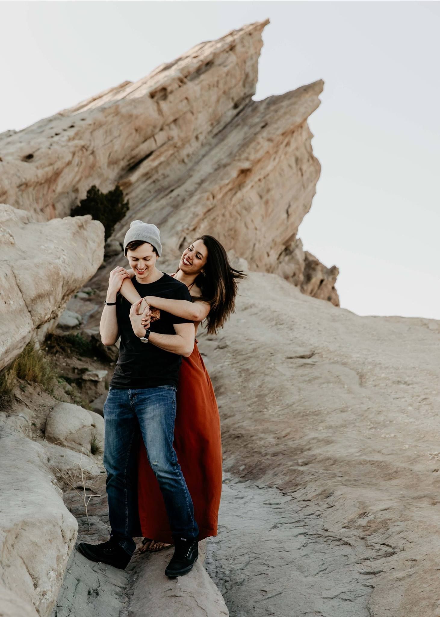 Laughing couple embracing on rocky desert terrain with large sandstone formations behind them.