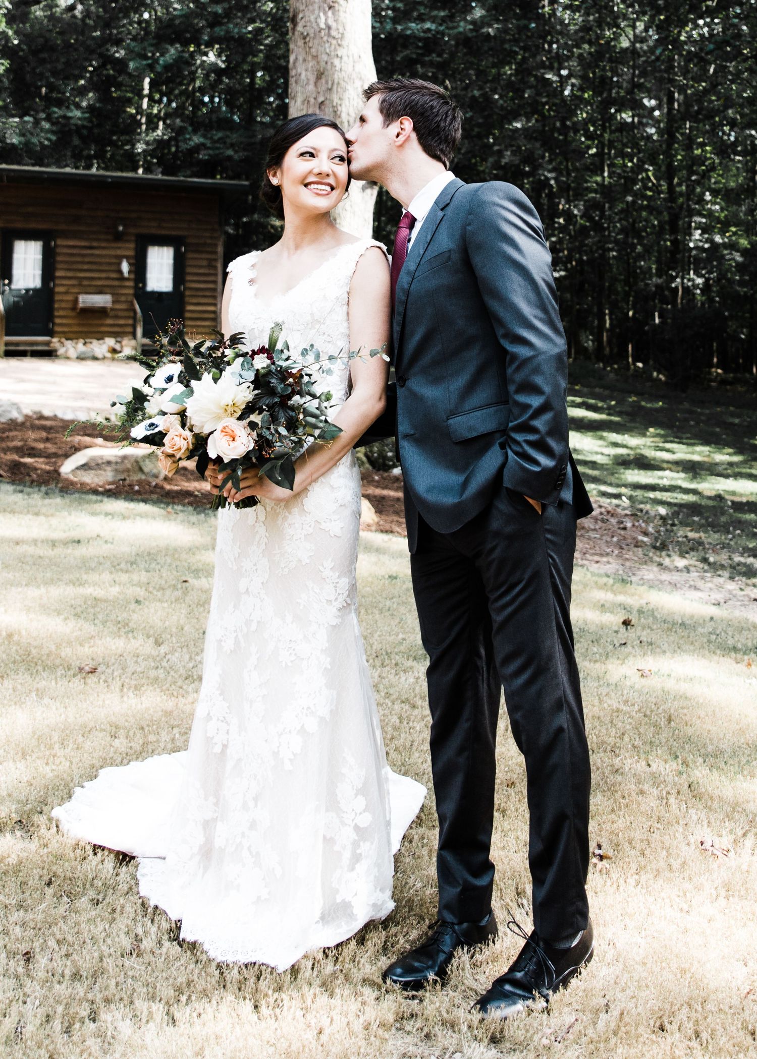 Groom kissing bride on cheek outdoors near log cabin, bride holding bouquet.