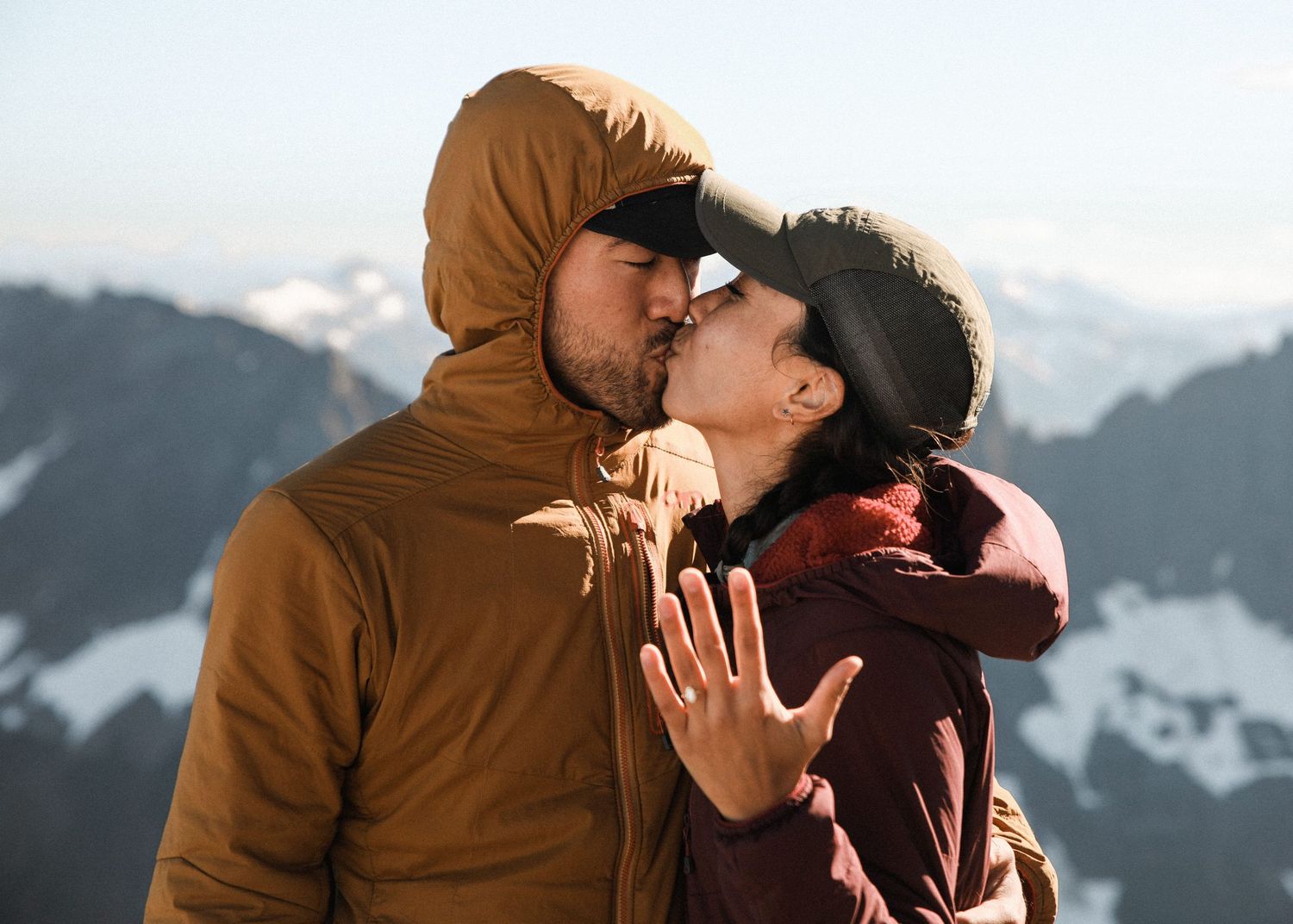 Engaged couple sharing a kiss on mountain summit while bride shows off new ring.