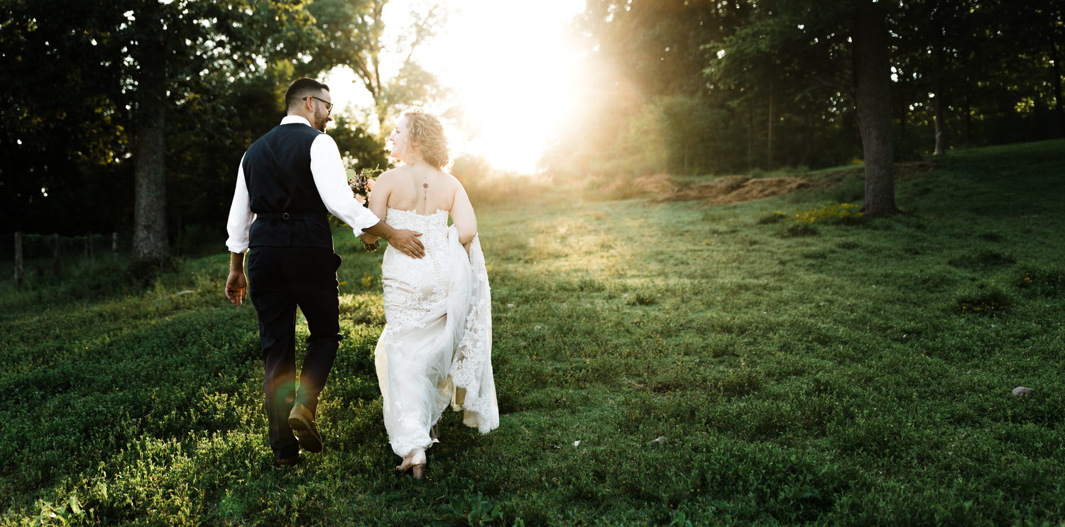 Bride and groom walking hand in hand through a sunlit green meadow surrounded by trees.