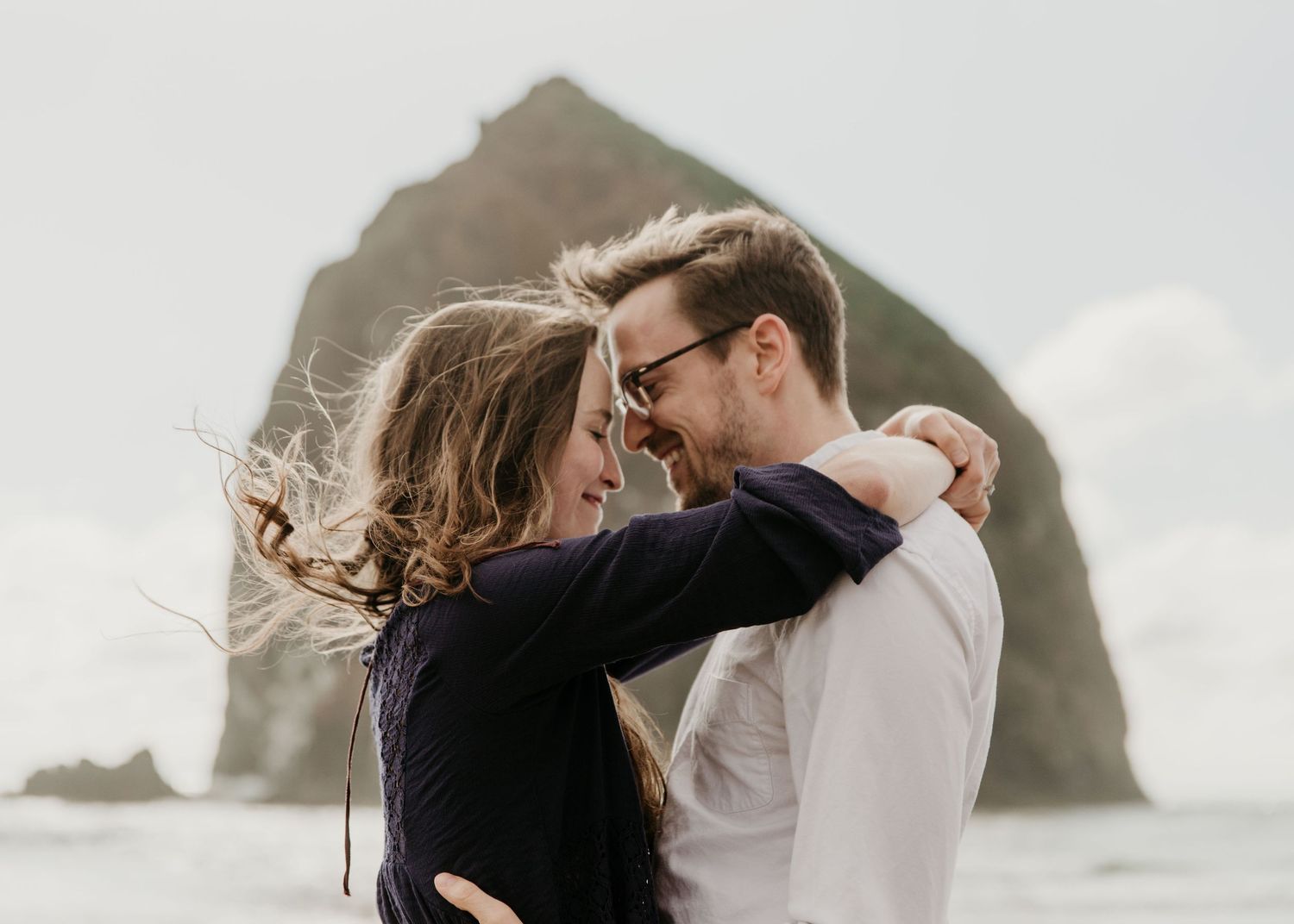 Romantic couple embracing and smiling on a windy beach with Haystack Rock in the background.