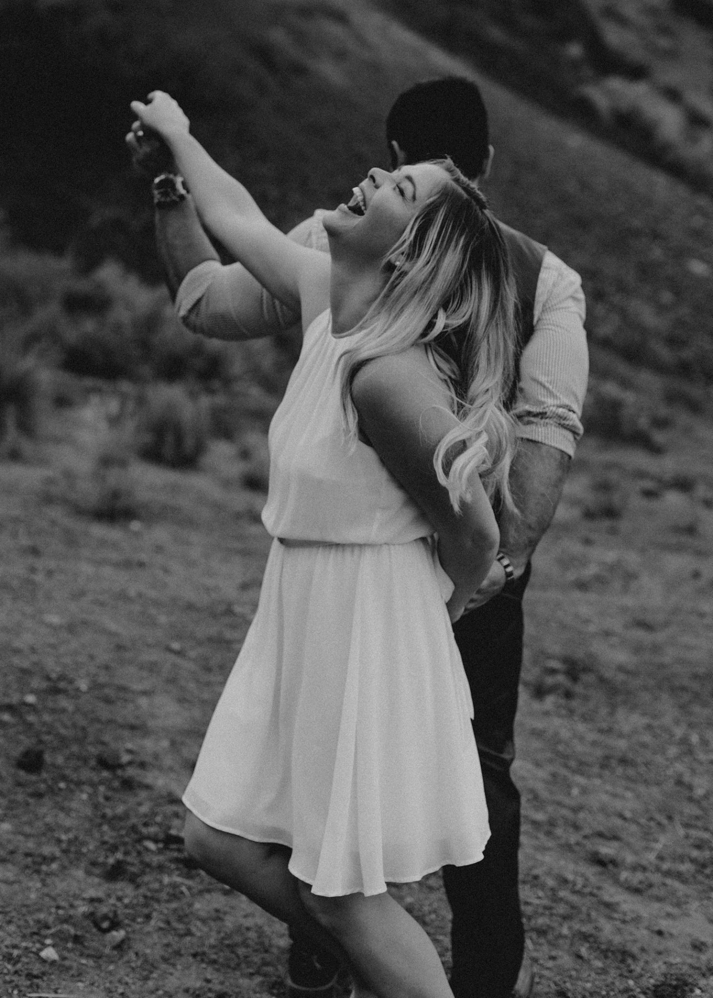 Black and white photo of joyful woman in white dress dancing with partner in scenic landscape.