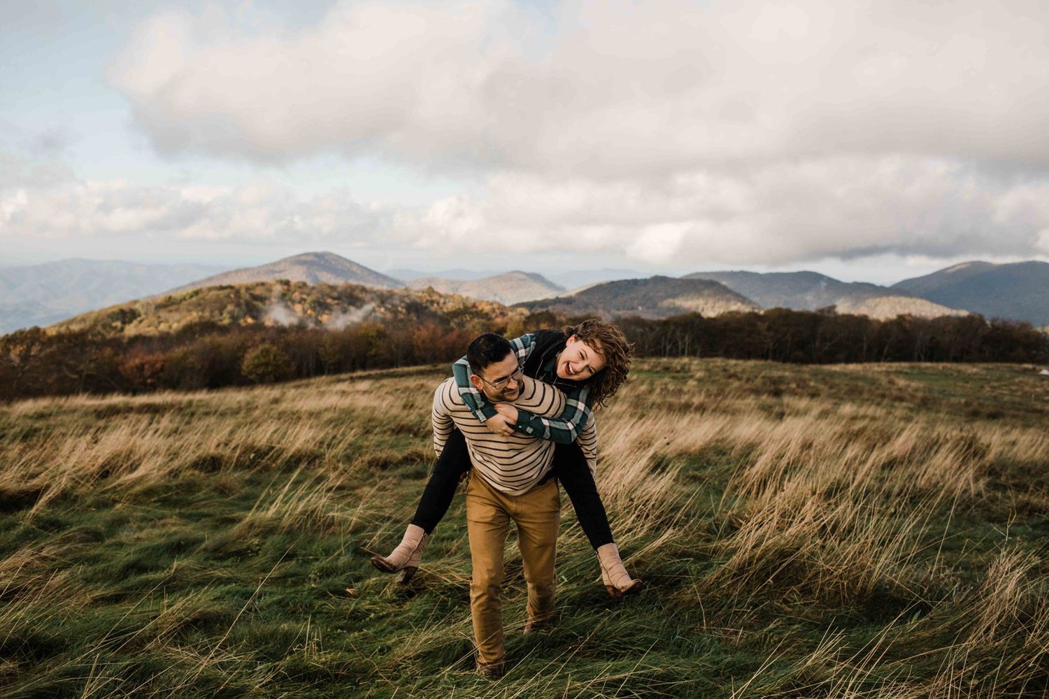 Happy couple laughing as man gives woman a piggyback ride in a scenic mountain meadow with tall grass.