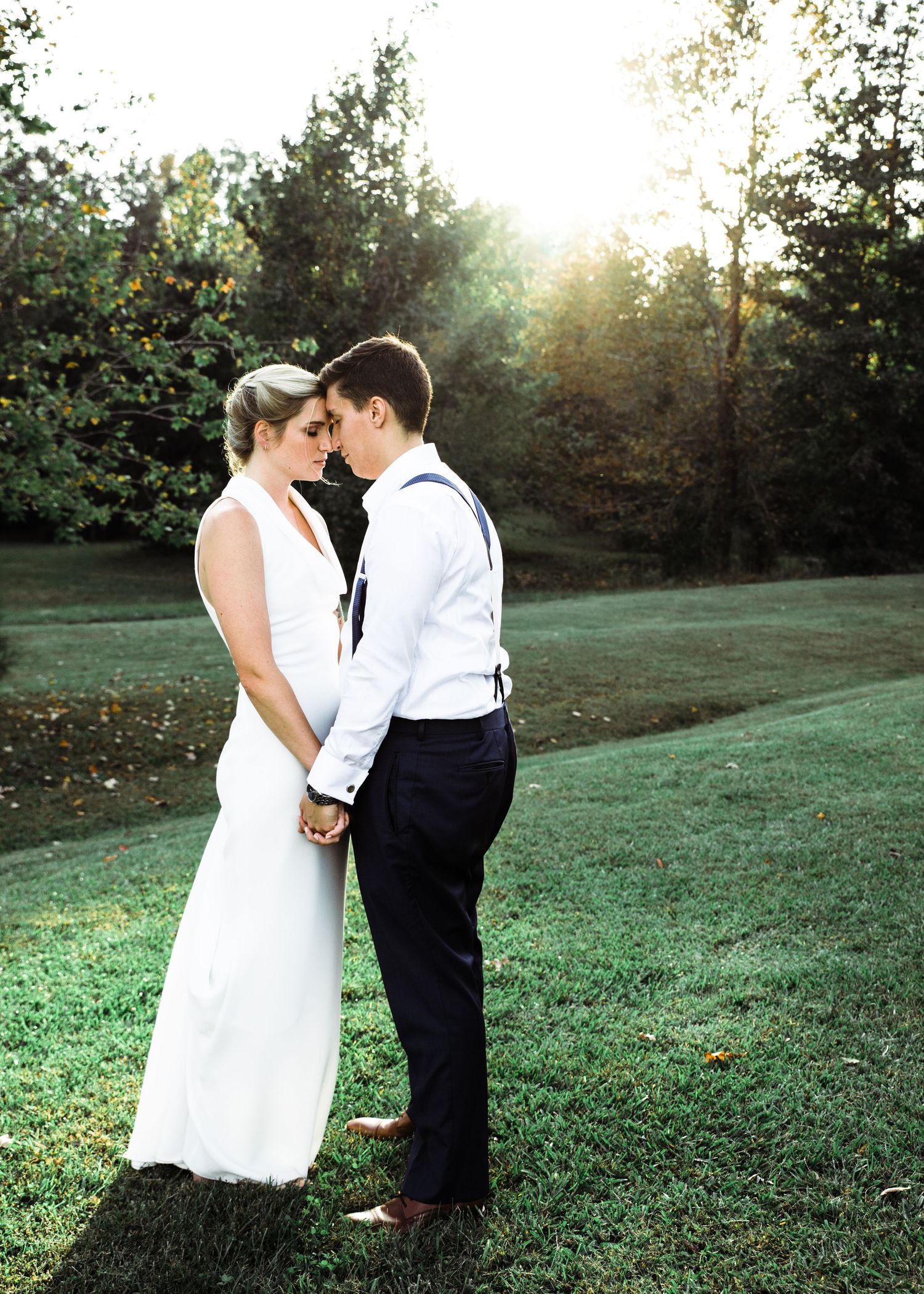 Bride and groom holding hands forehead to forehead in a sunlit green field surrounded by trees.