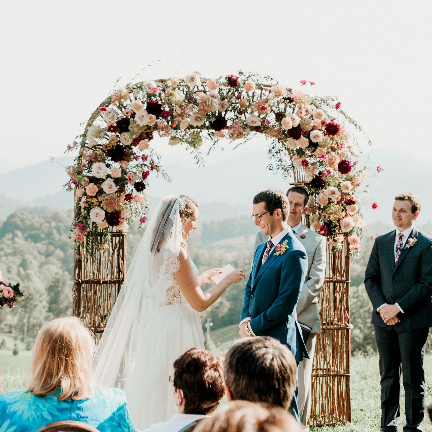 Outdoor wedding ceremony under a floral arch with bride and groom exchanging vows against a mountain backdrop.