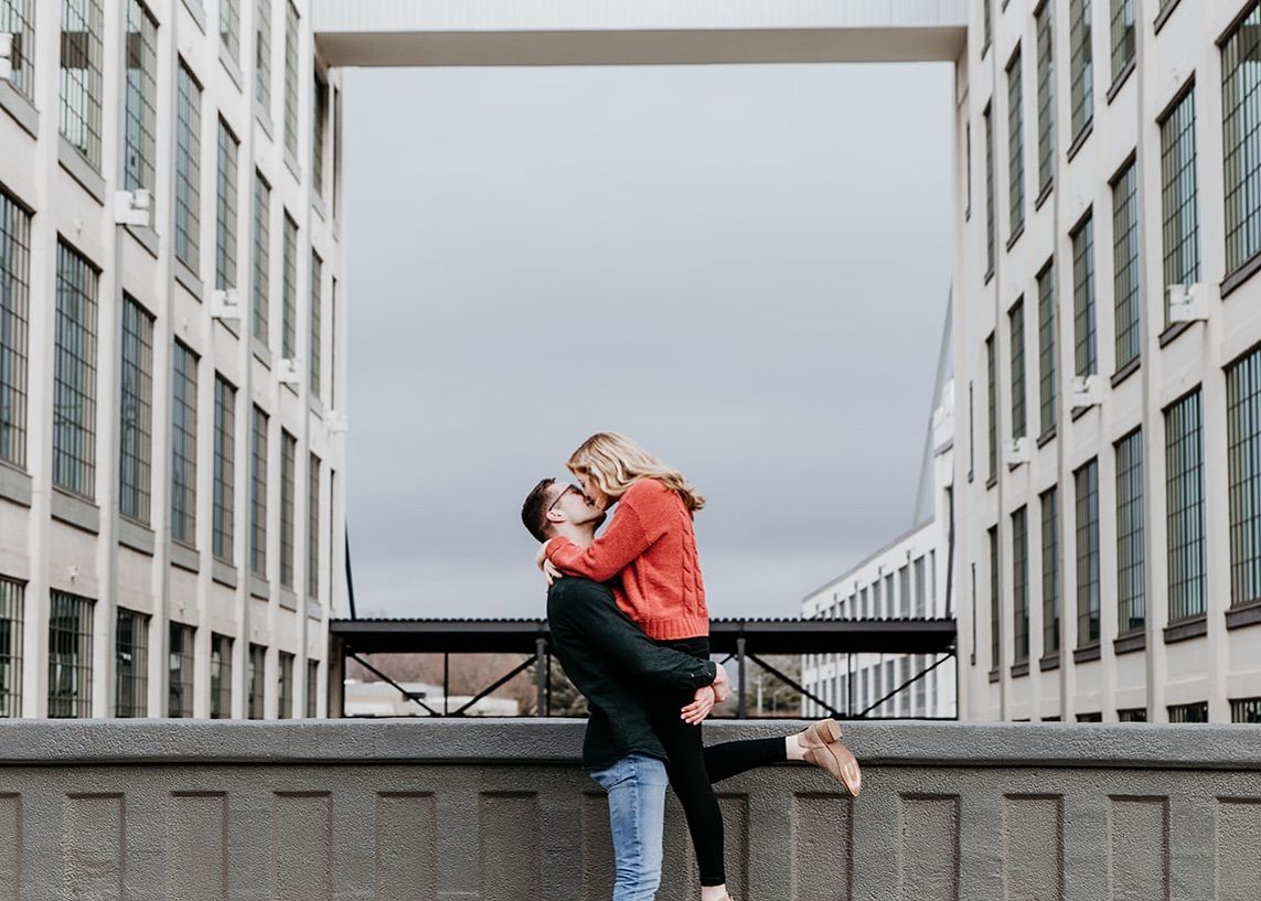 Man lifting woman in a romantic embrace between modern buildings in an urban outdoor setting.