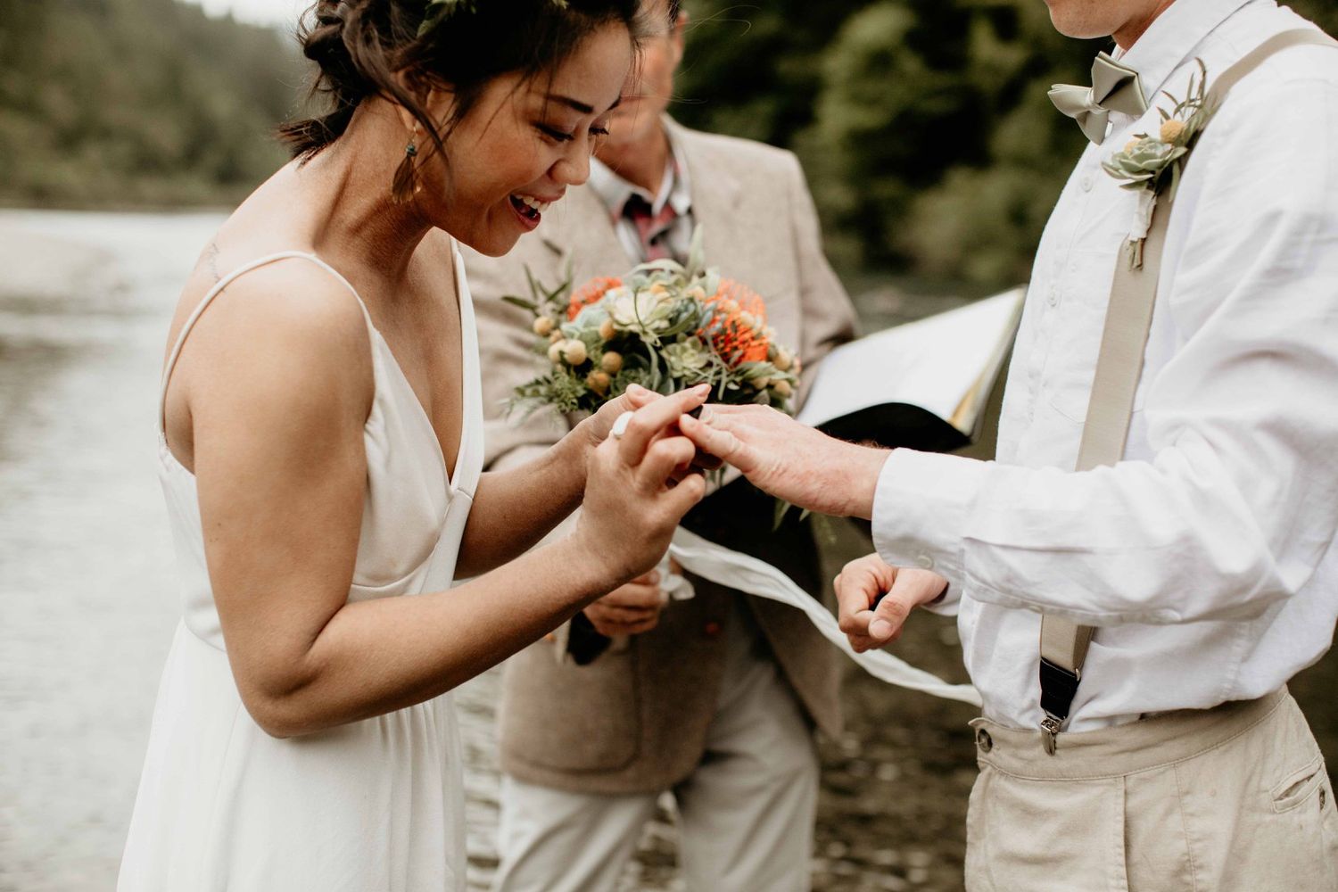 Emotional bride placing ring on groom's finger during intimate outdoor wedding ceremony by a river.