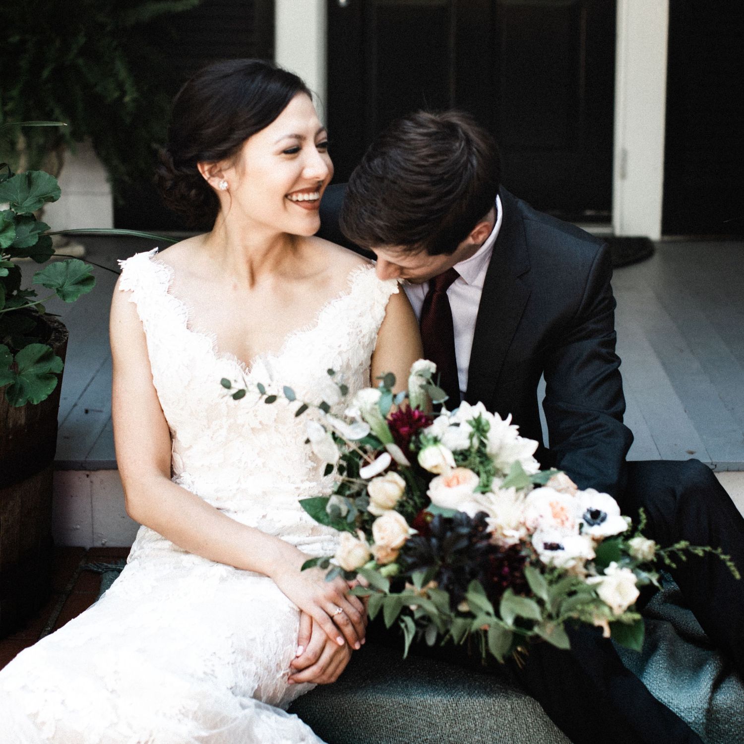 Smiling Asian bride in lace wedding gown holding bouquet while groom in black suit leans in close.
