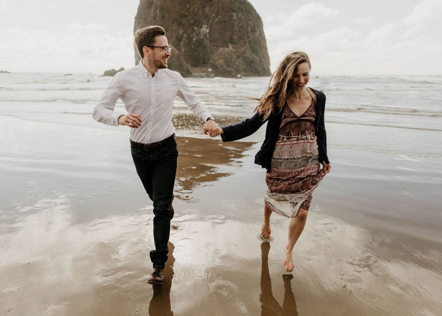 Couple running and holding hands on a wet beach with iconic Haystack Rock in the background.