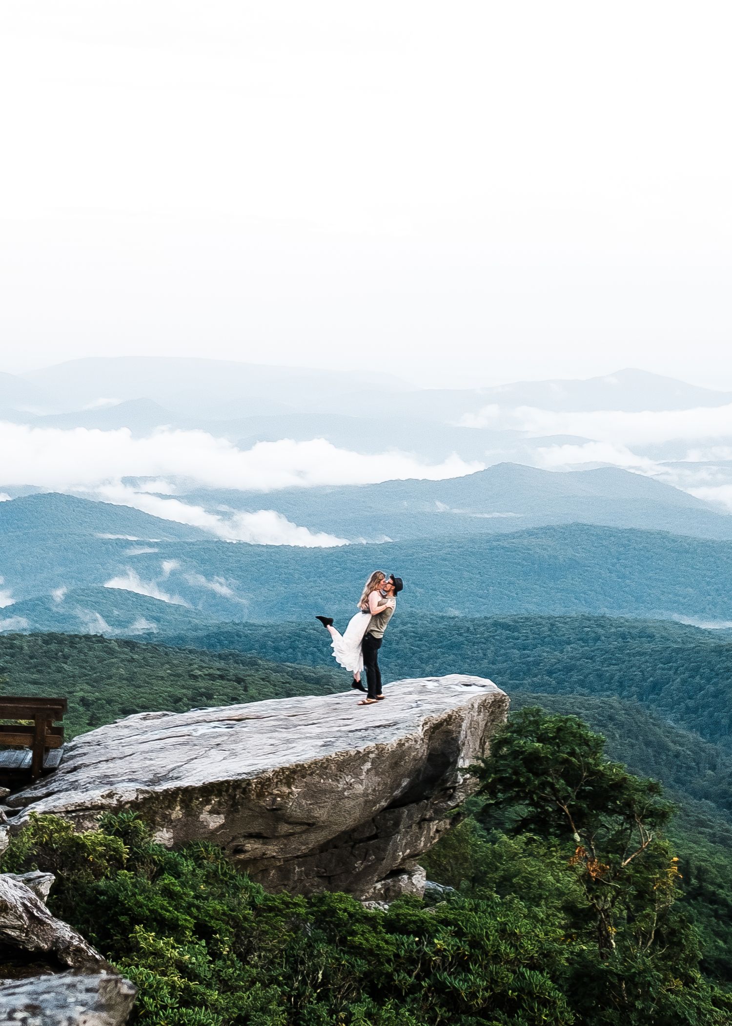 Couple embracing on a rocky mountain summit with sweeping misty blue ridge mountain views and lush green forest below.