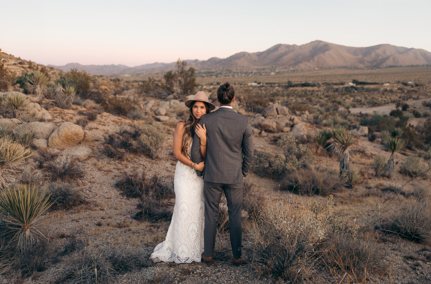 Bride and groom embracing in a desert landscape at sunset with mountains in the background.