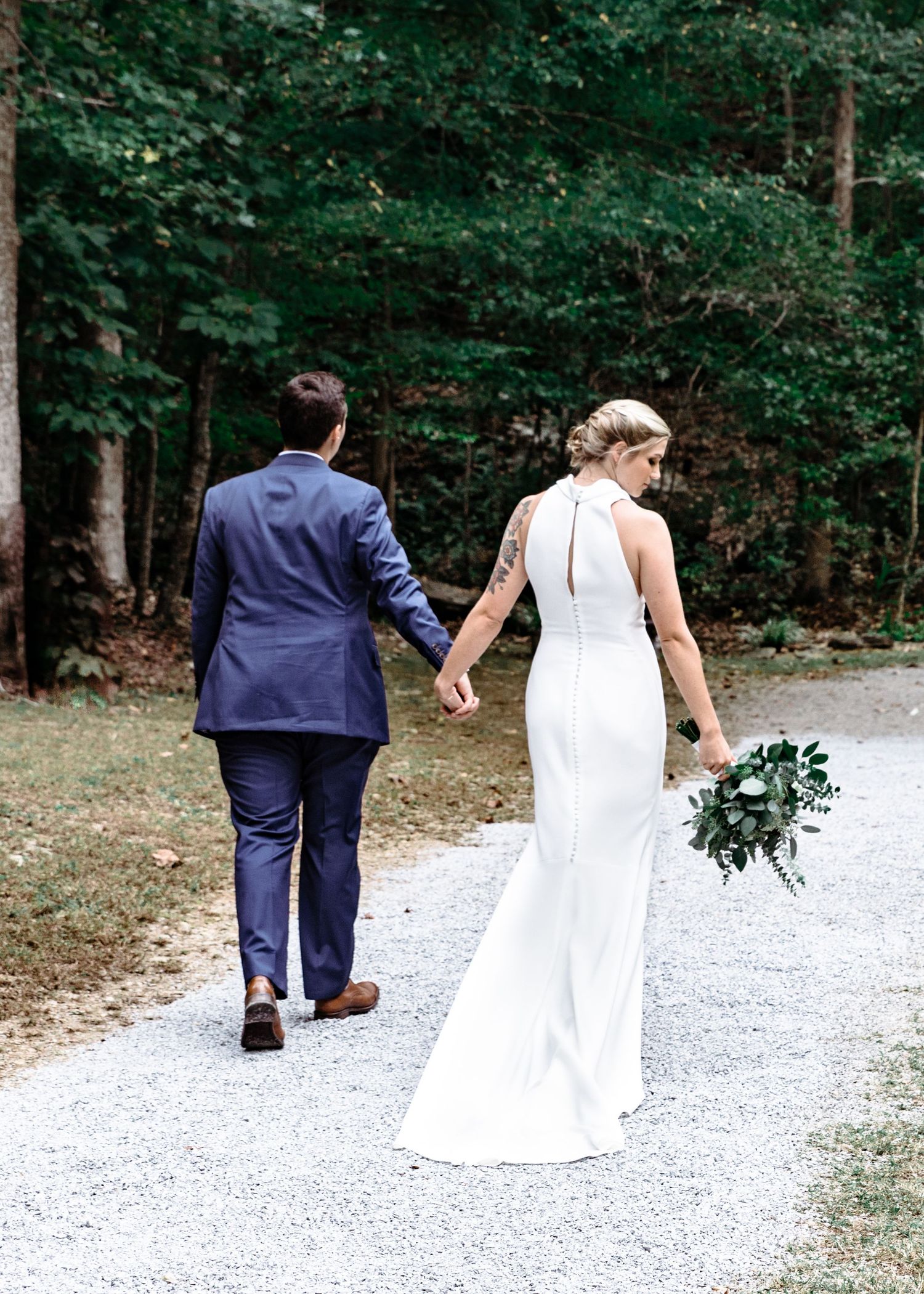 Bride and groom walking hand-in-hand down gravel path surrounded by lush green forest.