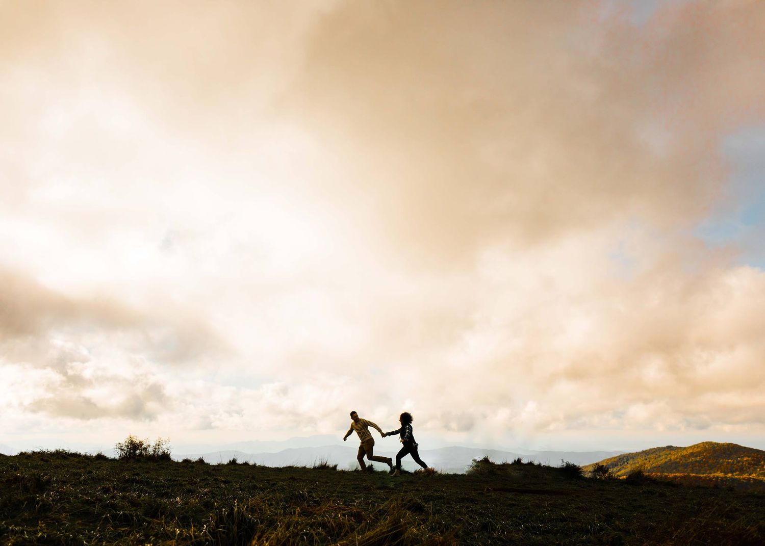 Silhouette of two people holding hands and running across a hilltop at golden sunset with dramatic clouds.