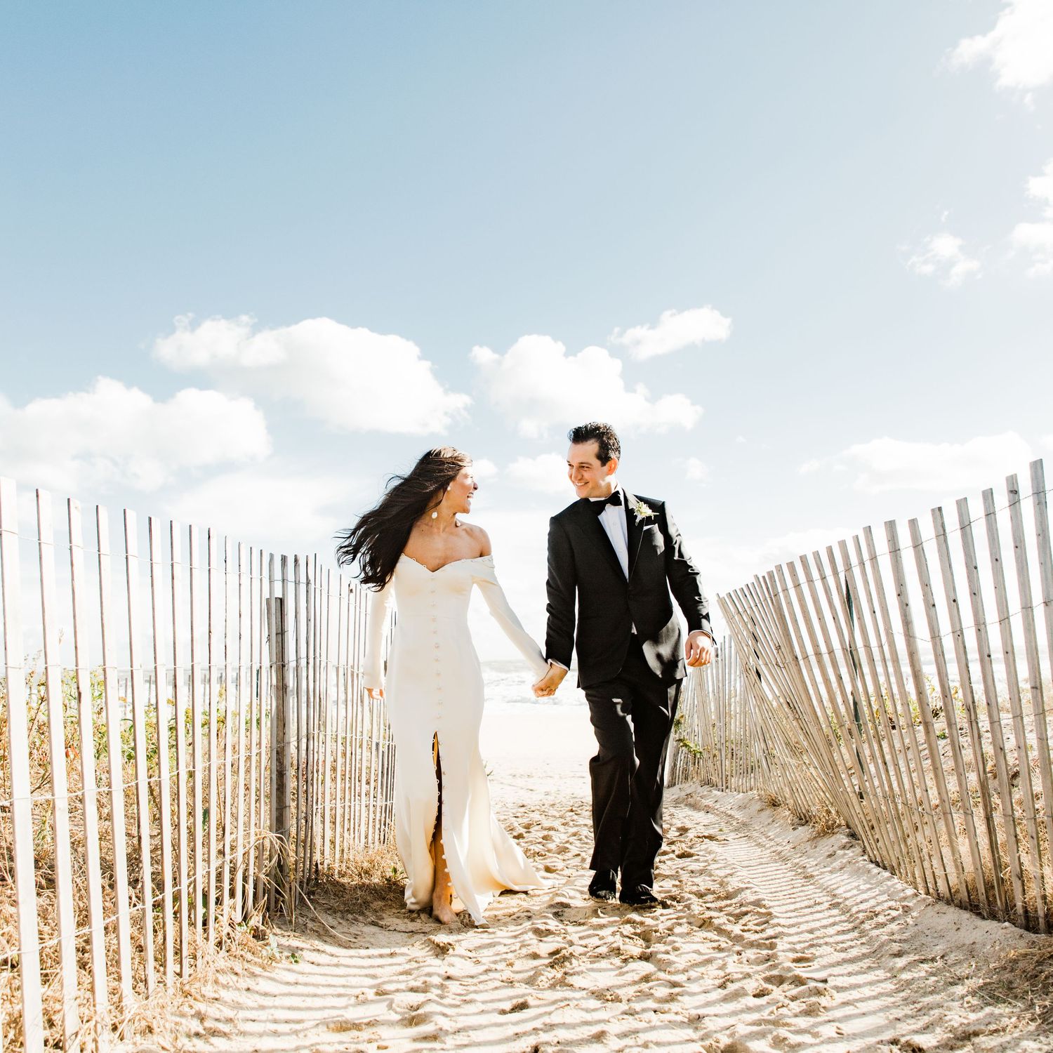 Bride and groom stroll together on a beautiful sandy beach path lined with rustic wooden fences.