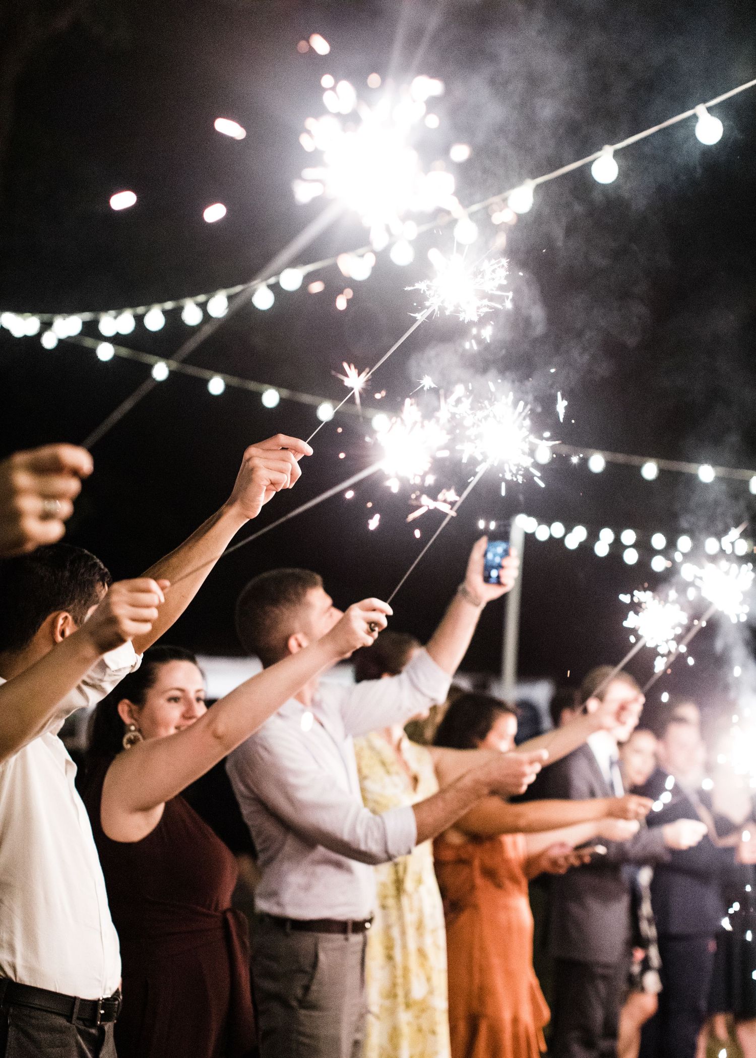 Wedding guests holding bright sparklers under string lights during an outdoor evening celebration send-off.