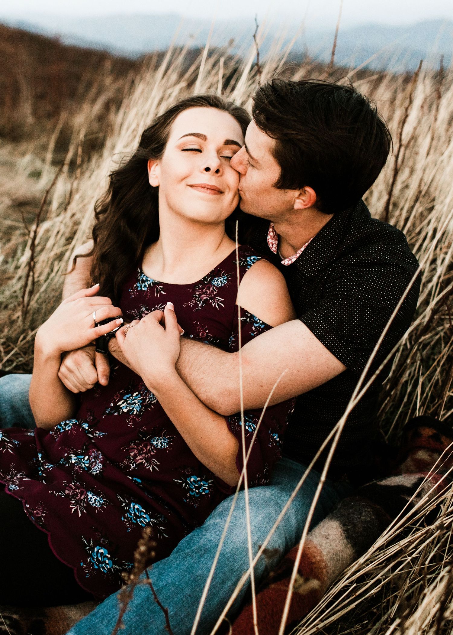 Couple sitting in tall grass, man kissing woman on cheek during romantic outdoor engagement photo session.