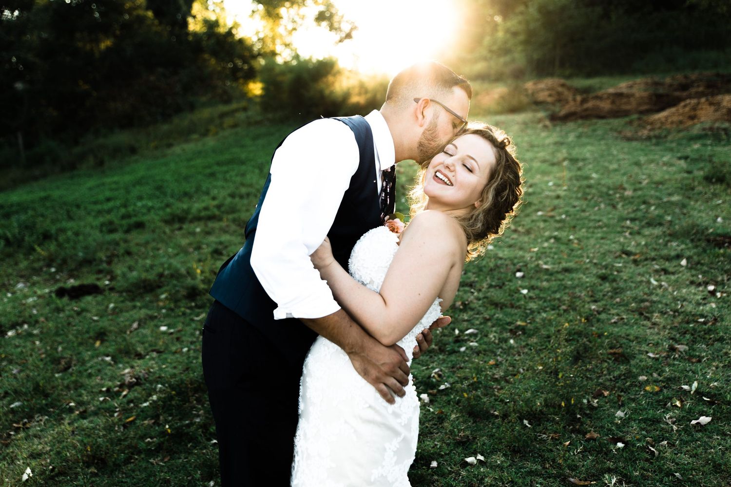 Groom dipping and kissing joyful bride in lace gown at sunset.