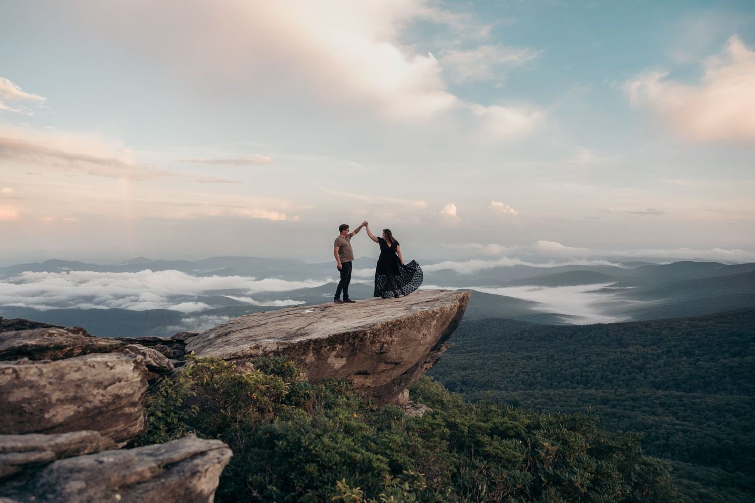 Couple holding hands on a dramatic rocky mountain overlook at sunset with misty valley below.