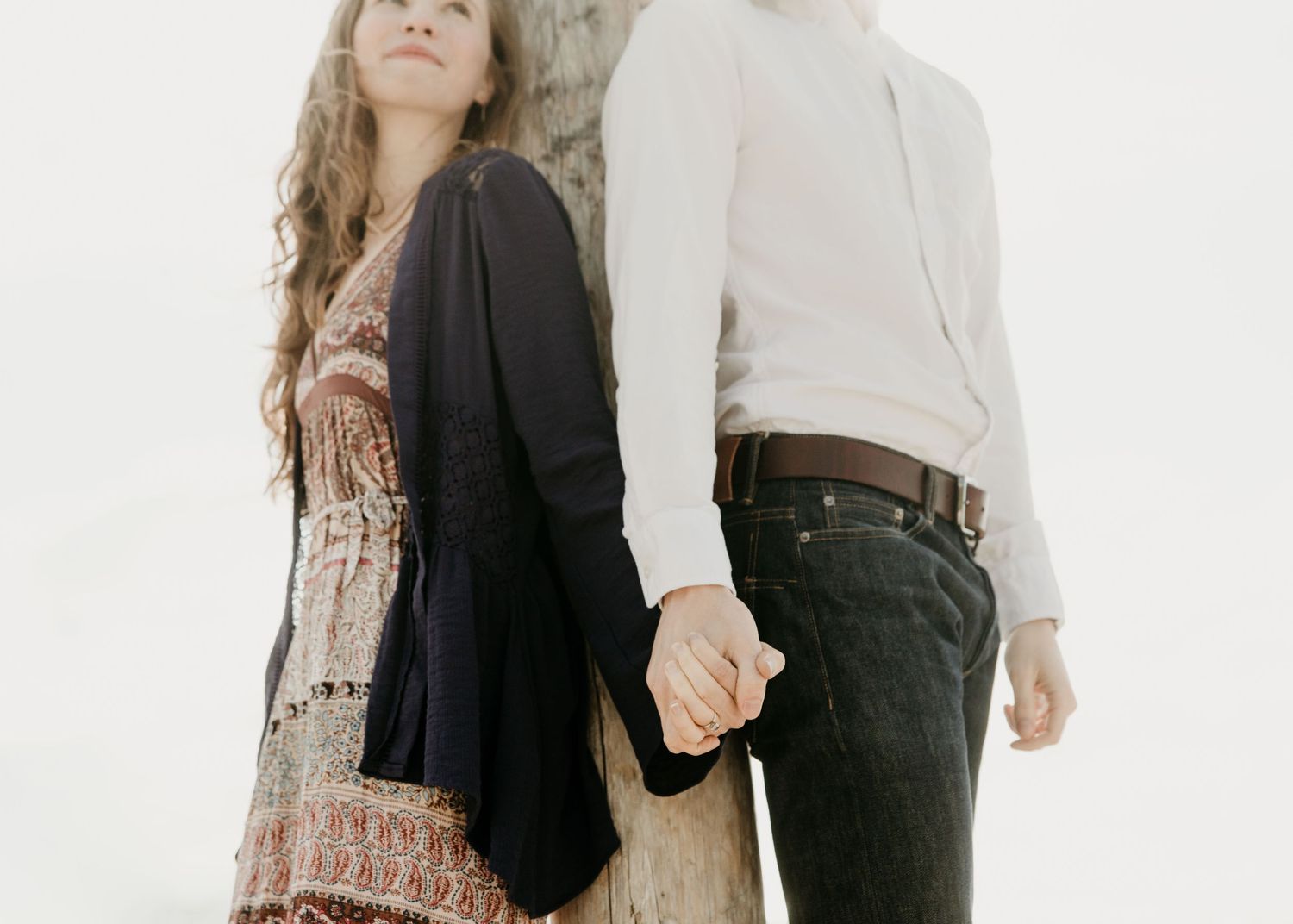 Young couple holding hands leaning against a tree in soft white light.