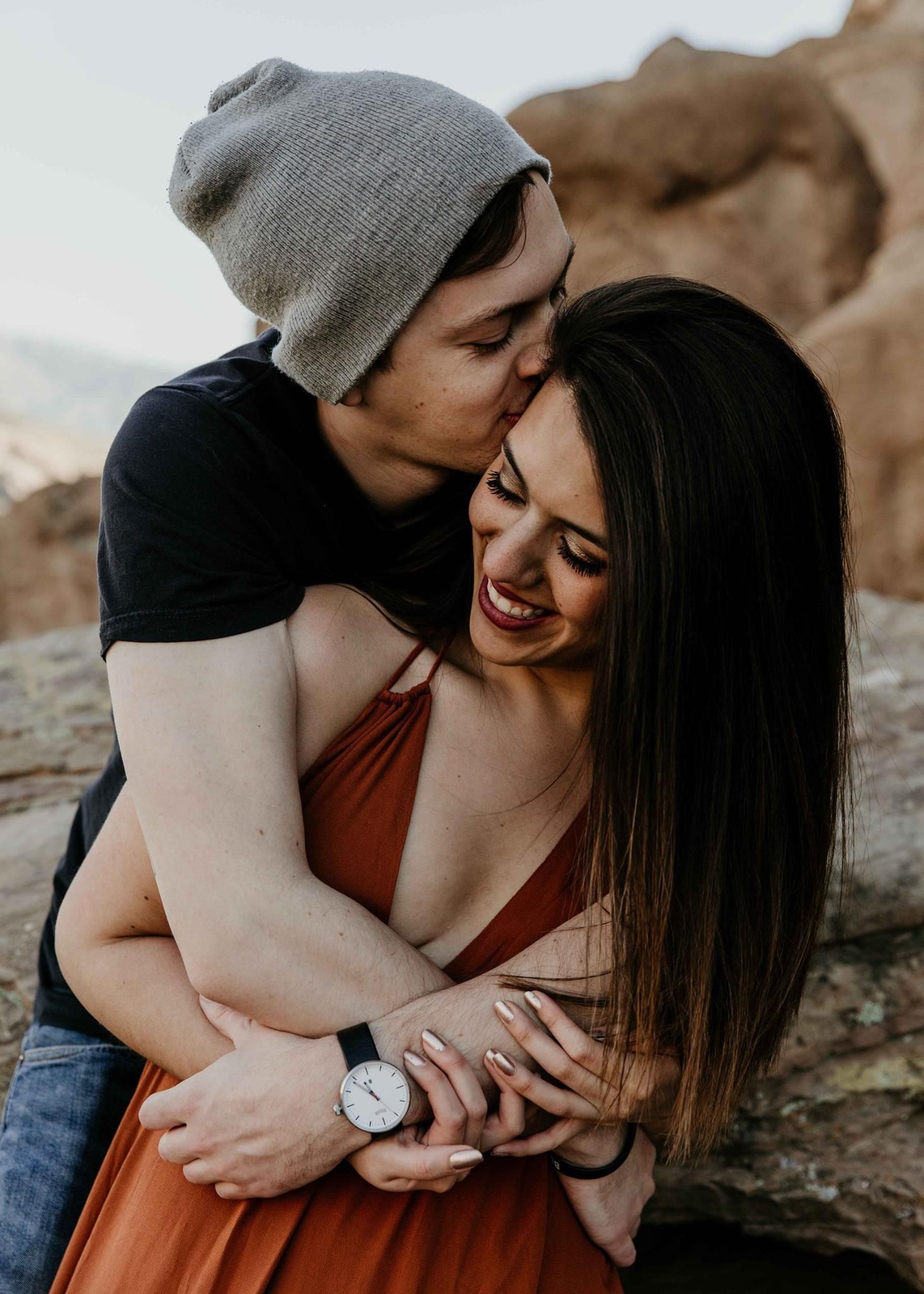 Man in gray beanie kissing laughing woman in rust dress during romantic outdoor engagement shoot.
