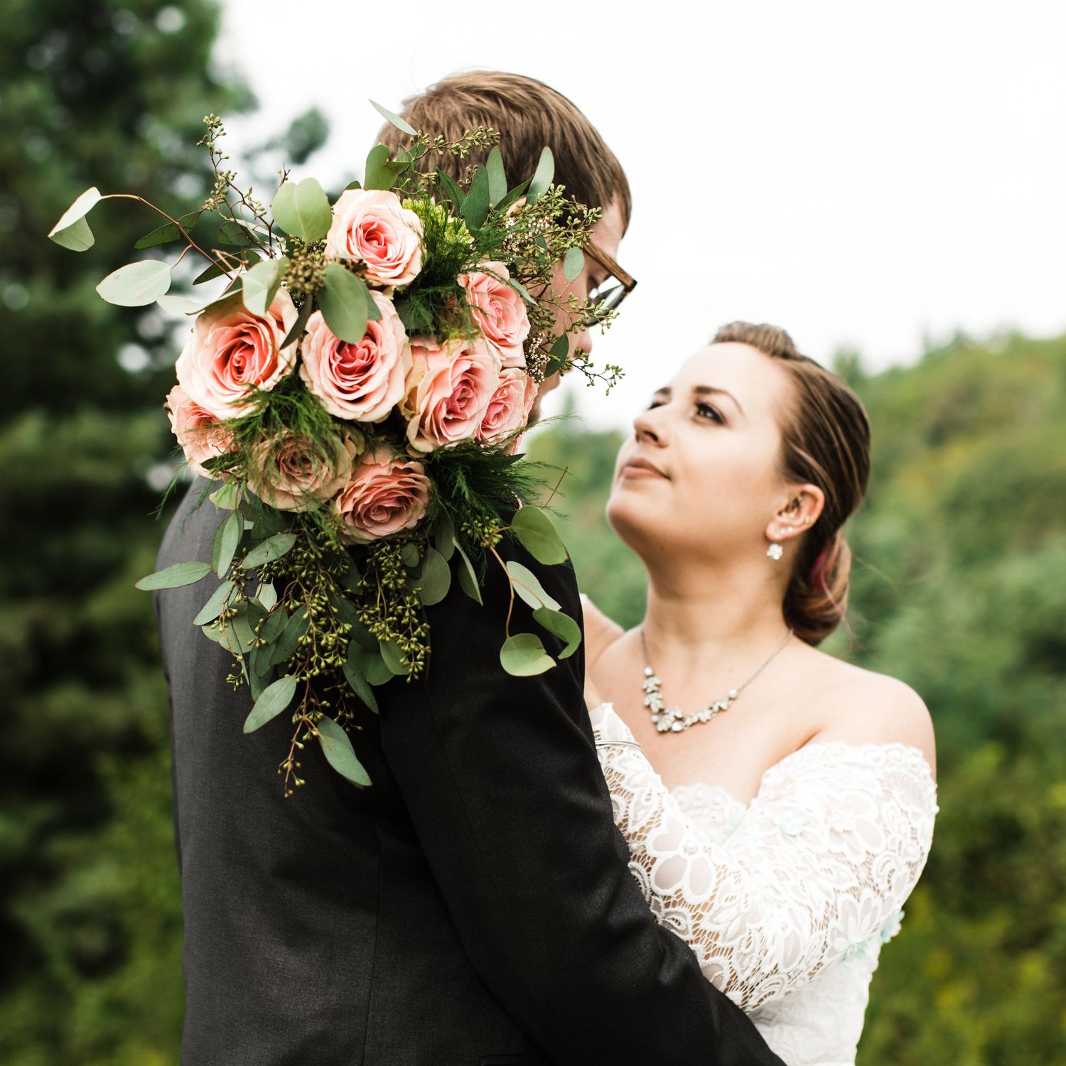 Bride in lace strapless gown leans against groom holding lush pink rose bouquet with greenery outdoors.